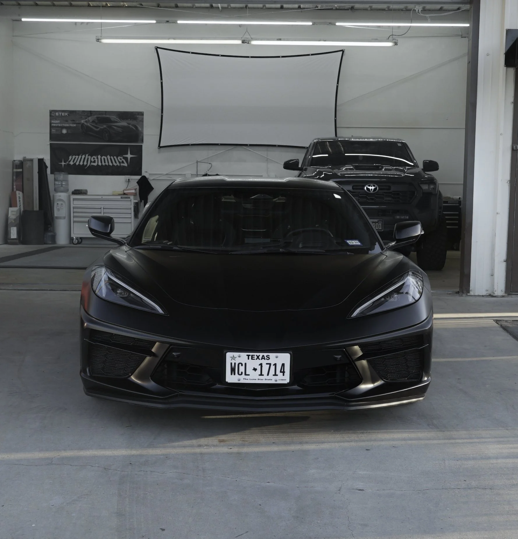A black sports car is parked inside a garage with a white backdrop and a Toyota vehicle behind it. The workshop area contains a tool chest, banners, and equipment. C8 Corvette Stingray wrapped in satin black colored paint protection film (colored PPF