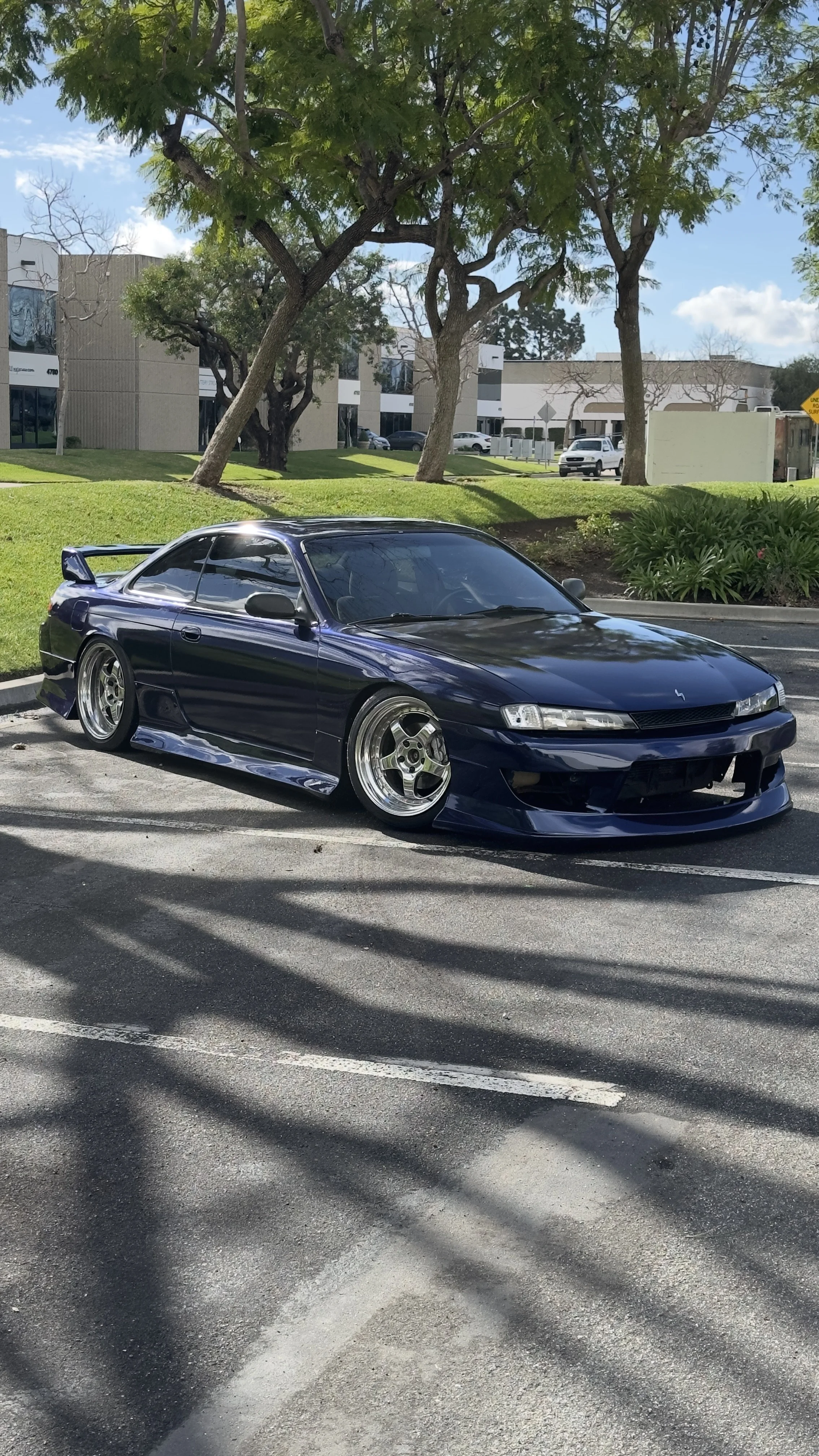 A dark blue sports car parked in an outdoor parking lot with trees and modern buildings in the background. S14 silva wrapped in gloss blue black iridescent vinyl wrap