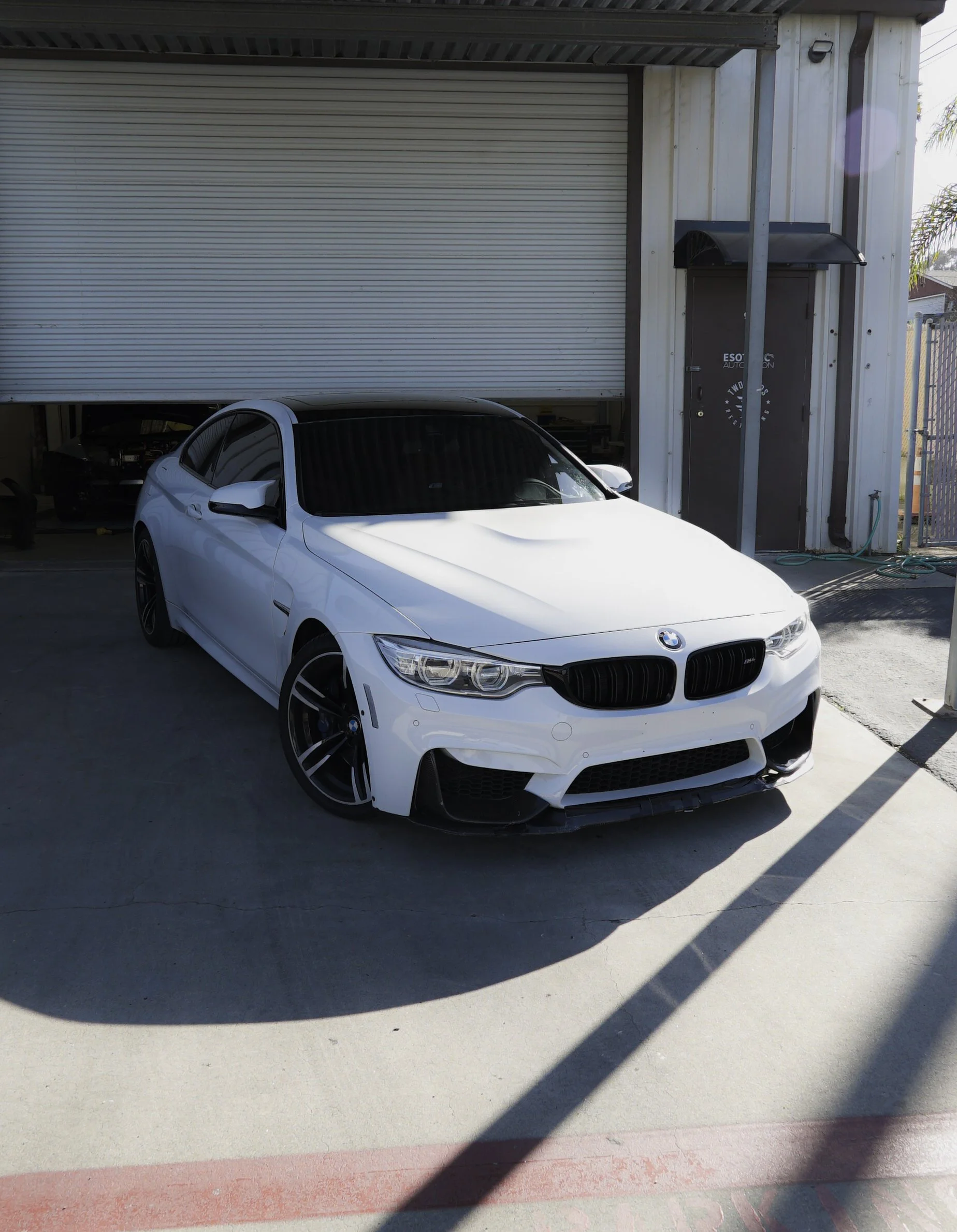A white BMW car parked in front of a garage with a partially closed roll-up door. BMW roof wrapped in gloss black ppf paint protection film