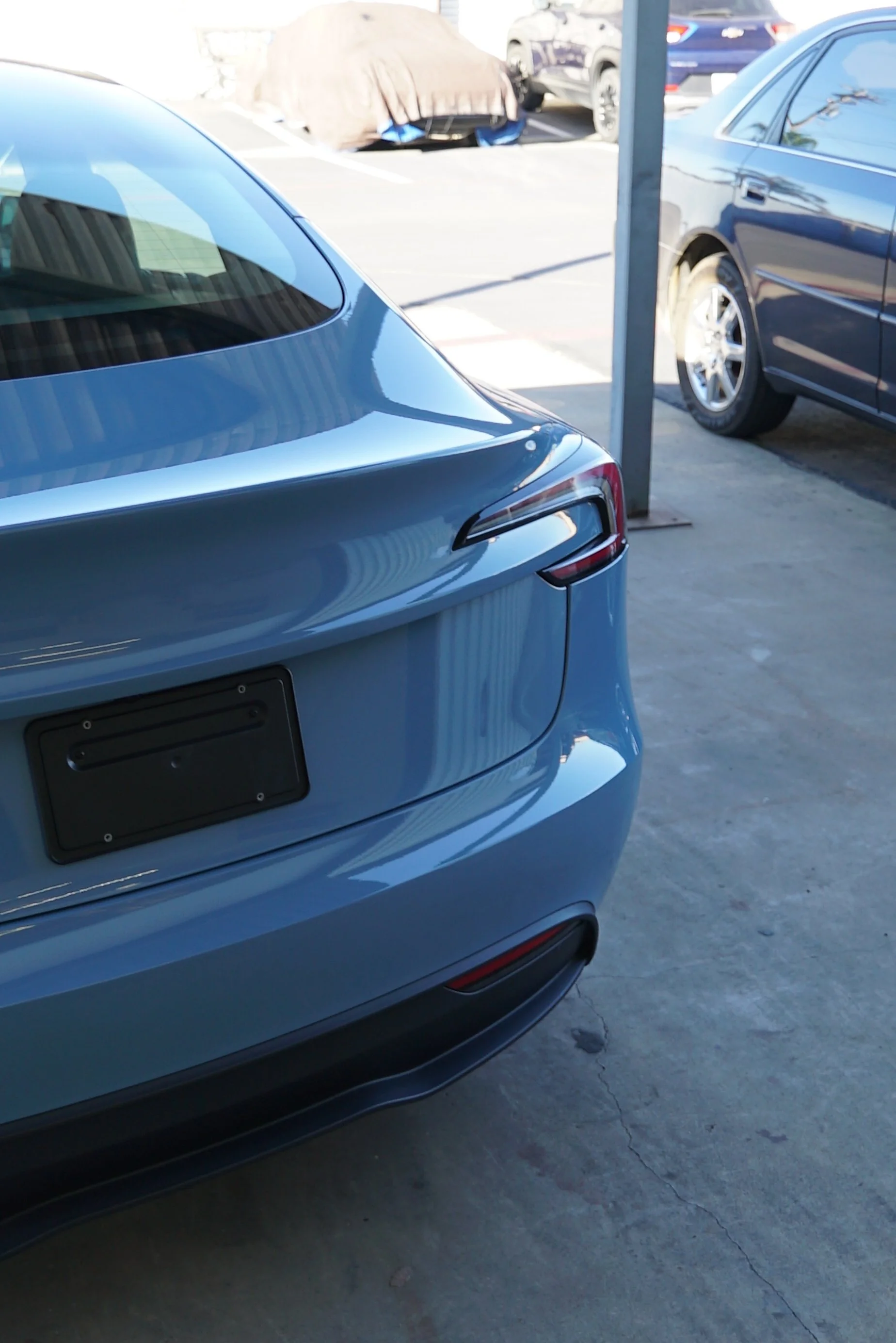 The rear end of a blue modern car parked in a lot, with part of a dark car and surroundings visible. Tesla is wrapped in colored paint protection film blue colored PPF