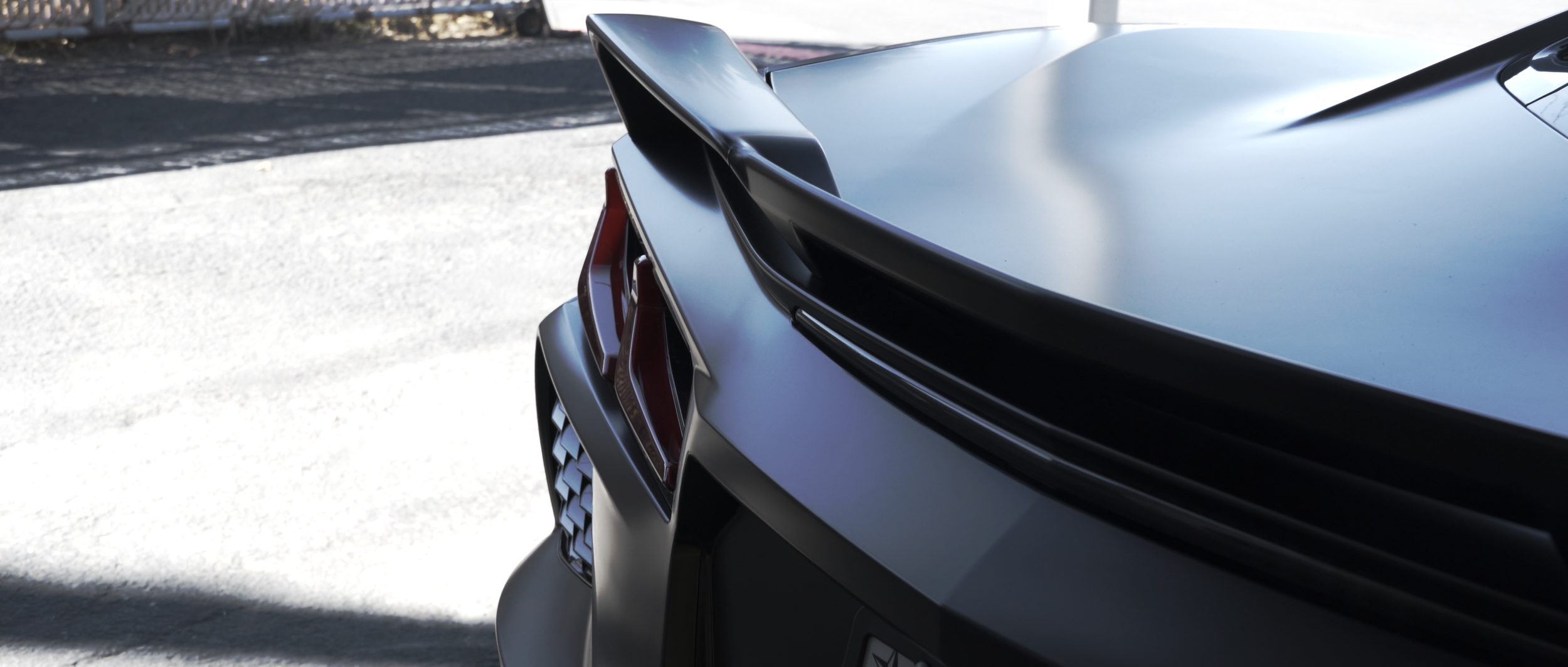 Close-up of the rear of a sleek, dark gray or black sports car with a prominent rear spoiler and distinctive taillights, parked outside on a bright day. C8 Corvette Stingray wrapped in satin black colored paint protection film (colored PPF)