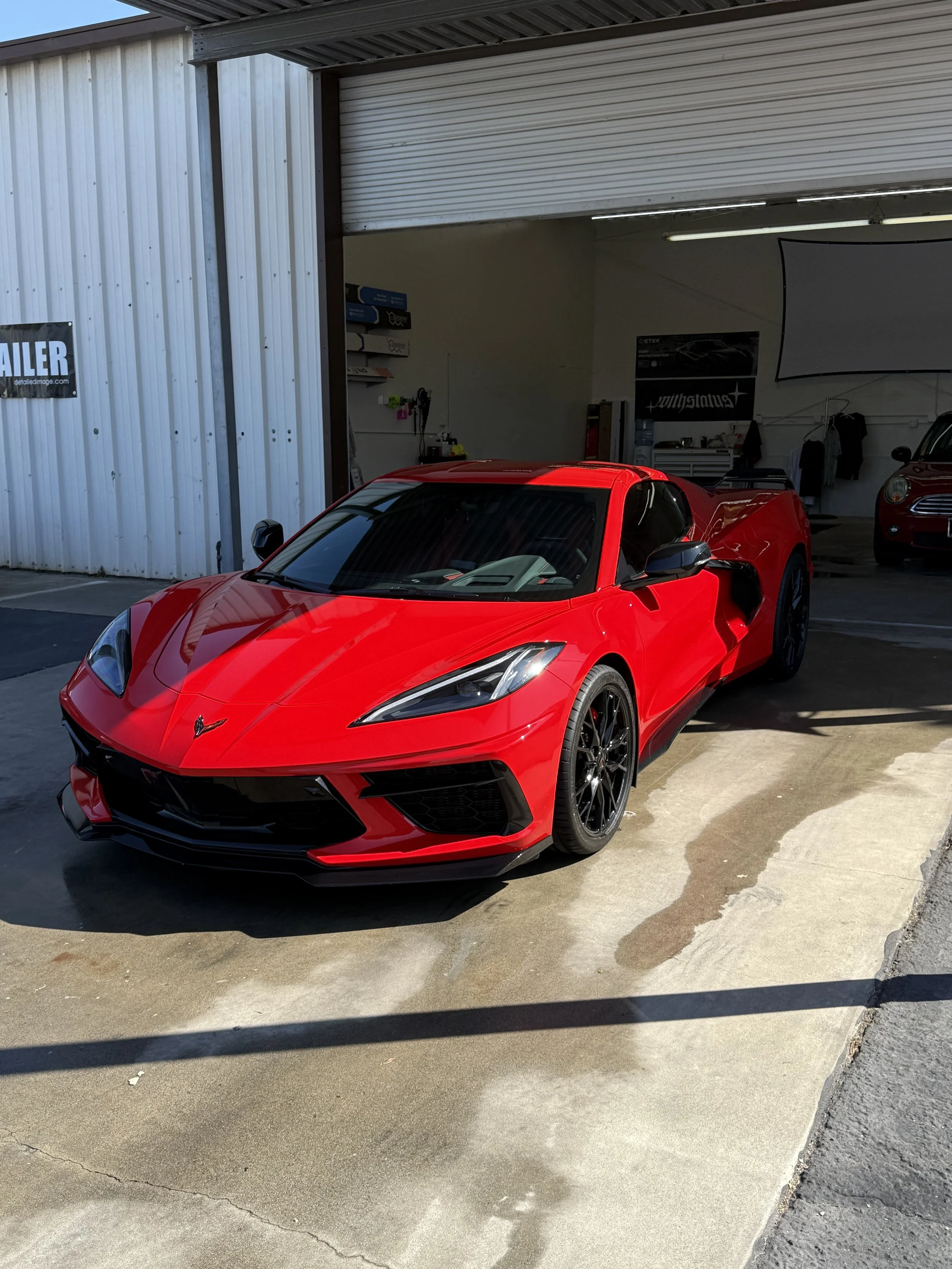 Red Chevrolet Corvette sports car parked outside a garage with open doors, with black wheels and tinted windows after gloss clear paint protection film was applied