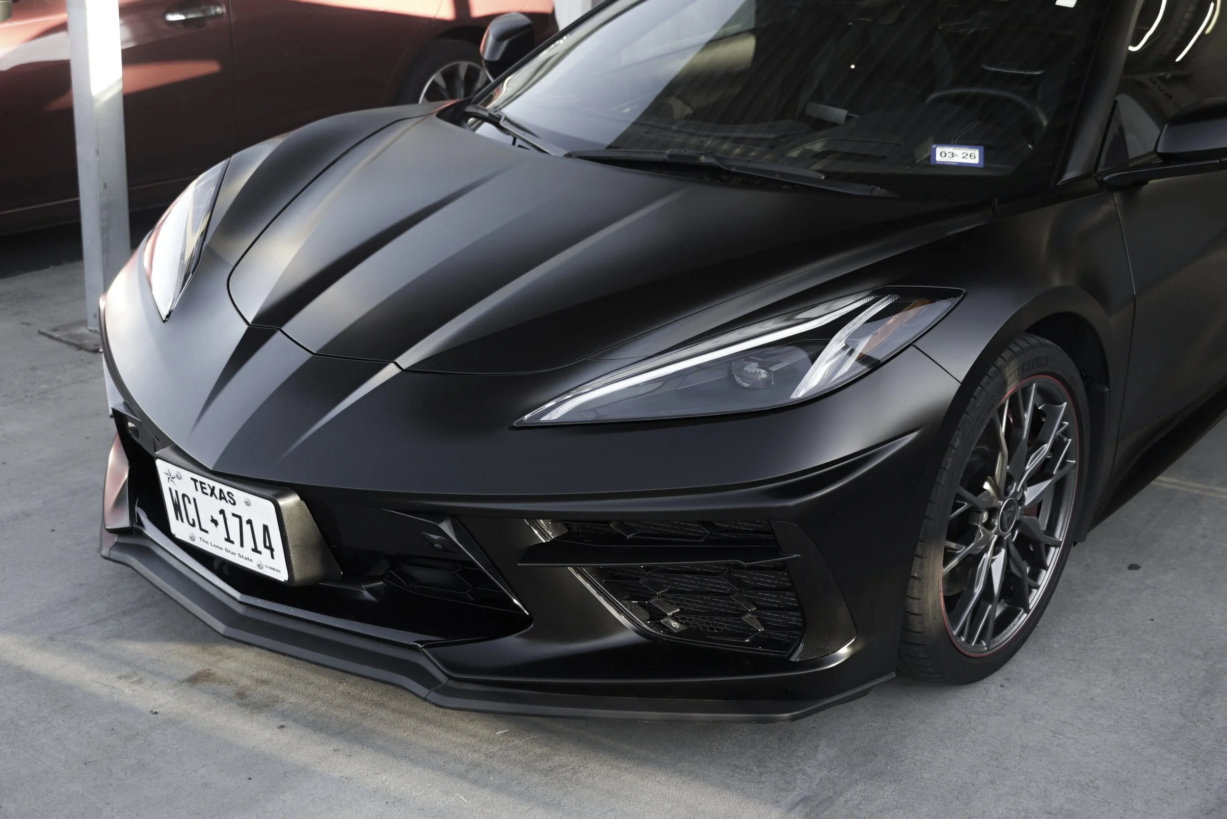 Close-up of the rear of a sleek, dark gray or black sports car with a prominent rear spoiler and distinctive taillights, parked outside on a bright day. C8 Corvette Stingray wrapped in satin black colored paint protection film (colored PPF)