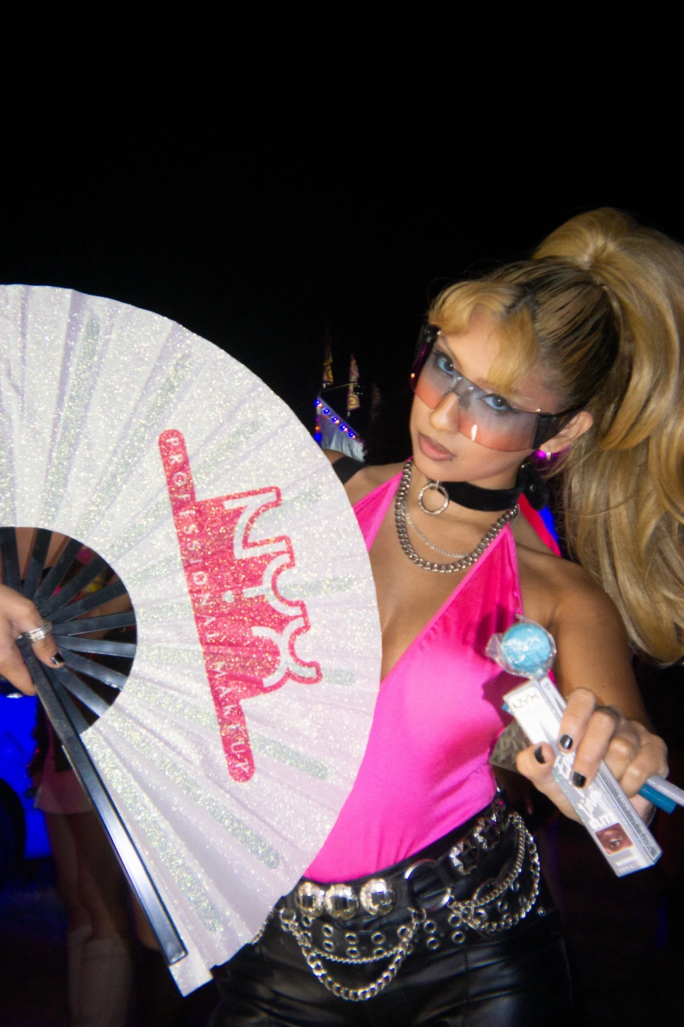 Woman in pink top and black leather pants holding a white glittery fan with pink logo, wearing large hair in pigtails, sunglasses, jewelry, and holding a lollipop, at night.