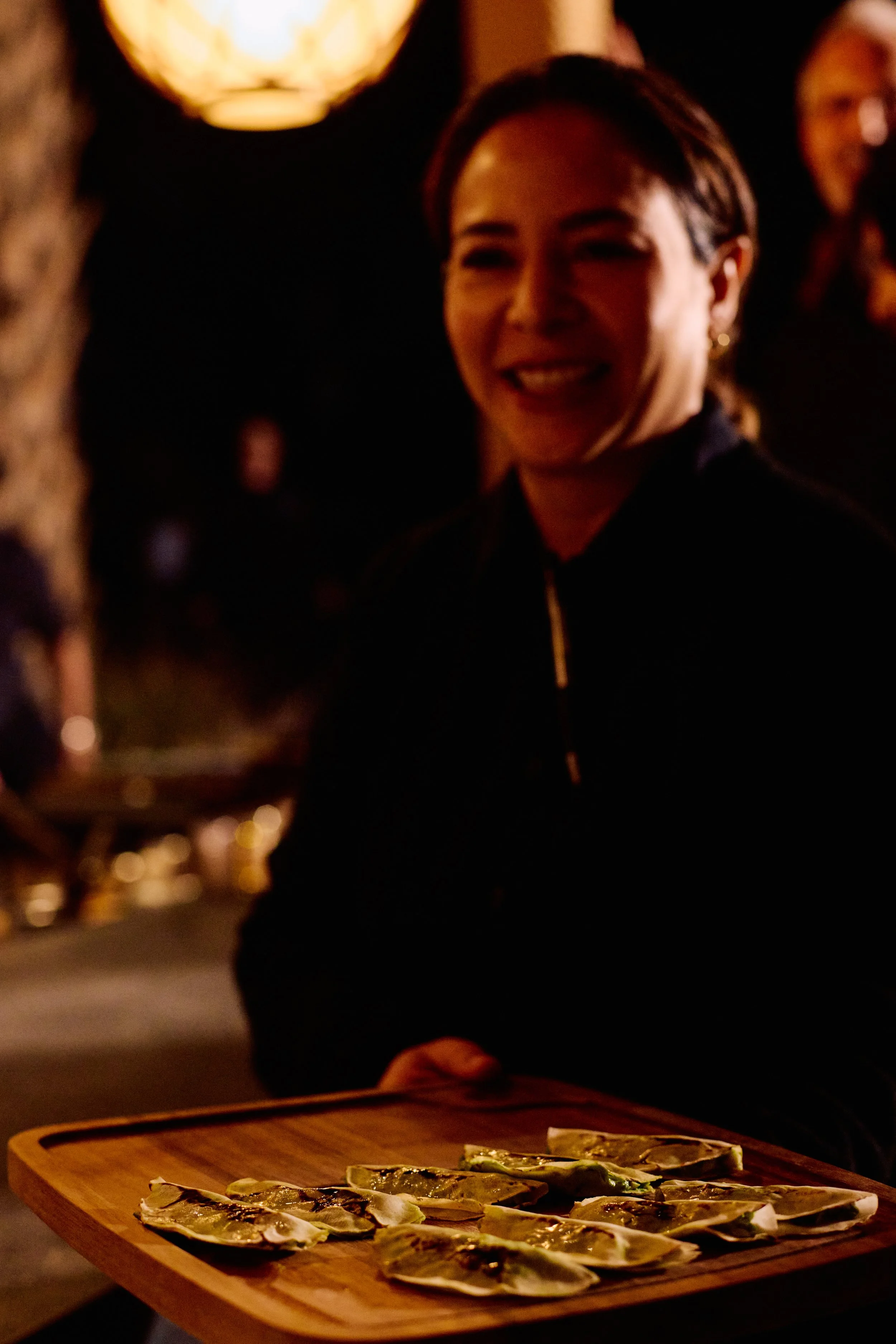 Woman smiling at a dimly lit outdoor restaurant holding a wooden tray with oysters on it.