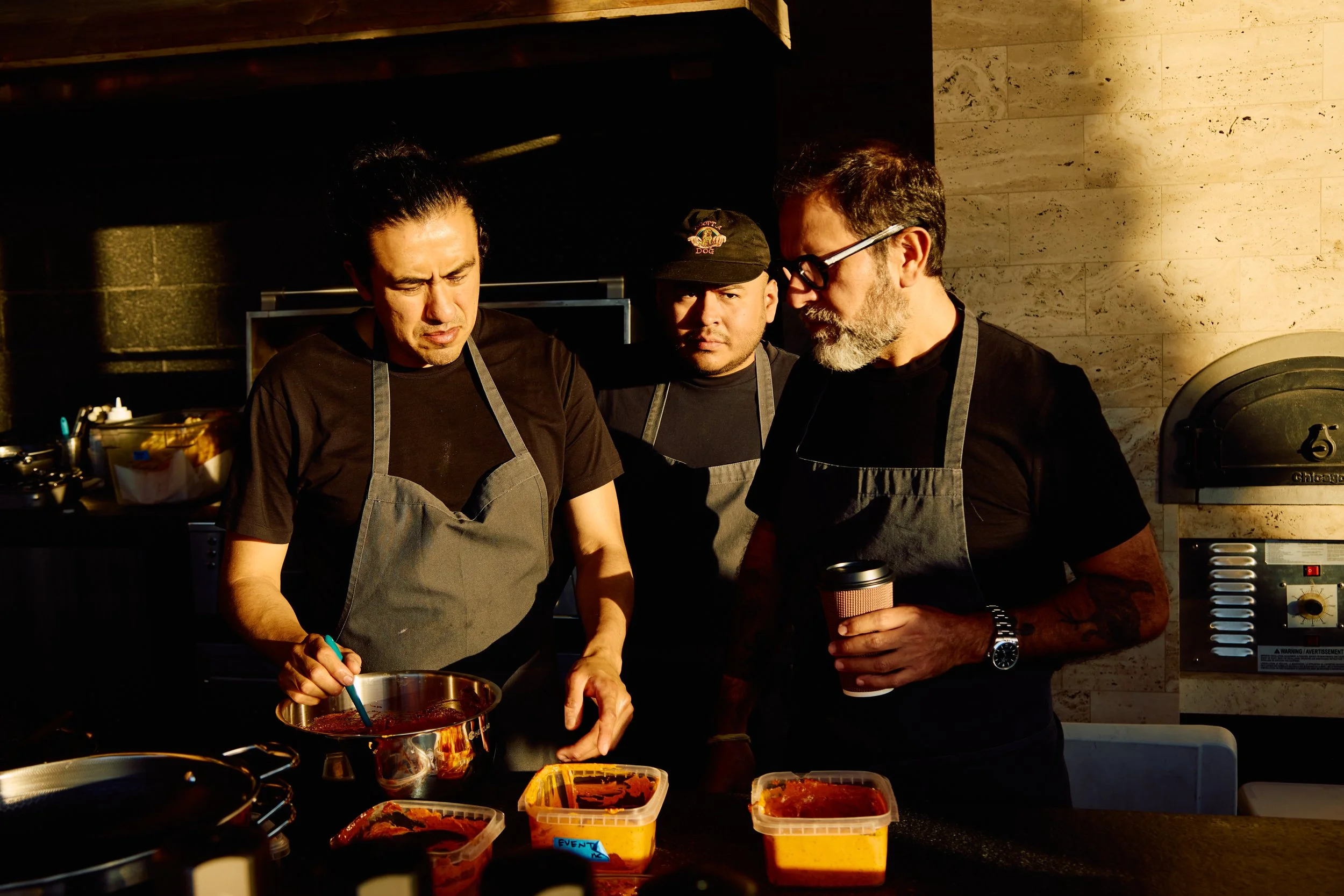 Three men in black shirts and gray aprons cooking together in a modern kitchen, one stirring a pot, another holding a coffee cup, with containers of sauce on the counter.
