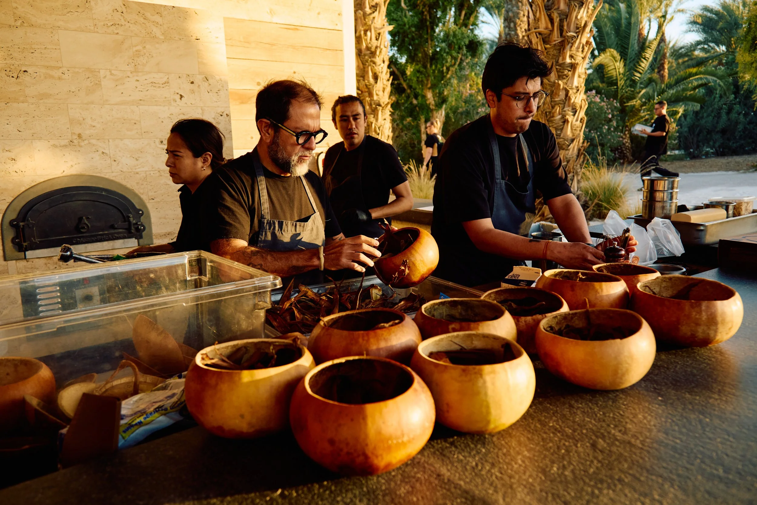 Four people preparing food outdoors with multiple wooden bowls on a counter and palm trees in the background.