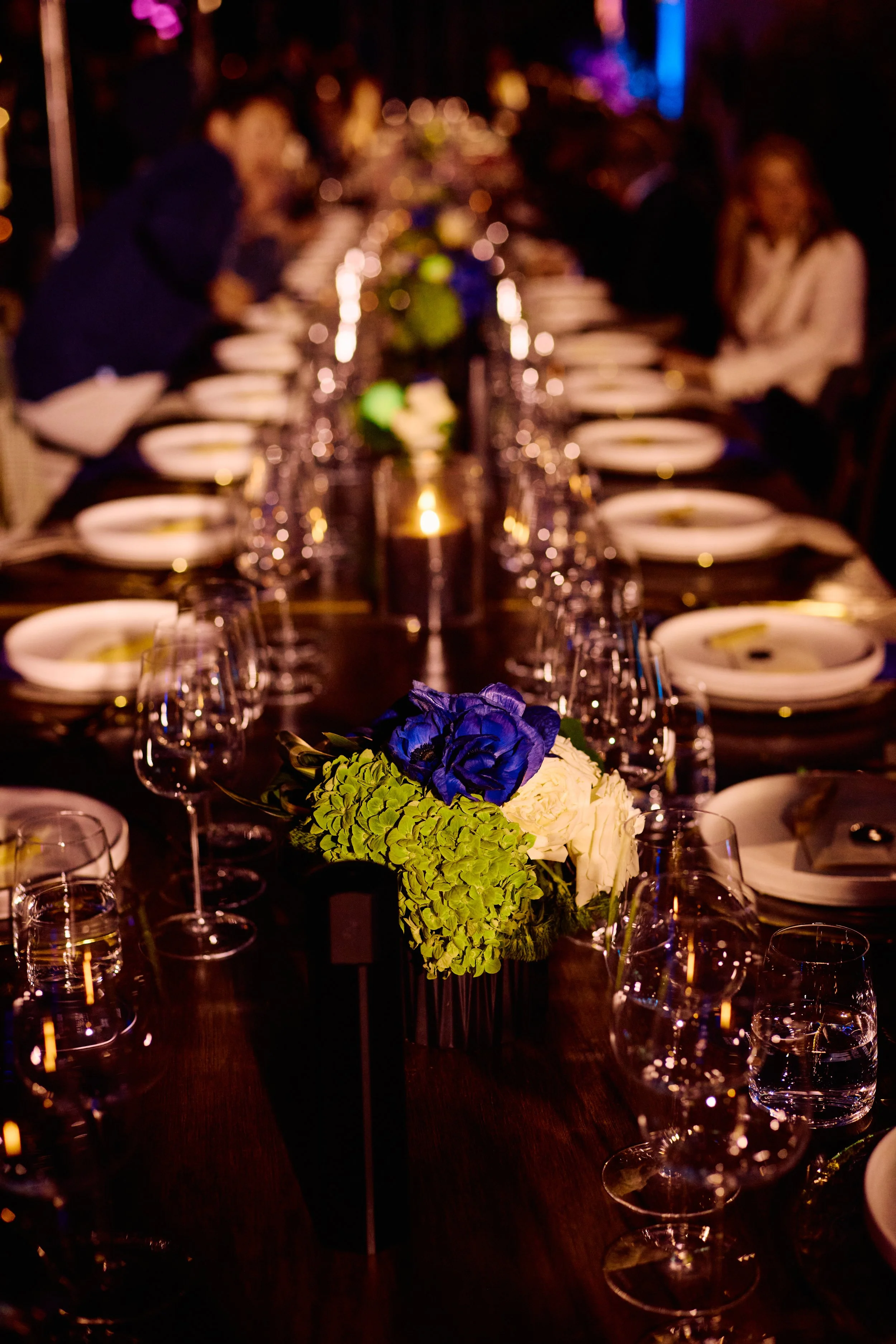 A long dining table set for a formal event with flower arrangements and glassware, with blurred guests in background.