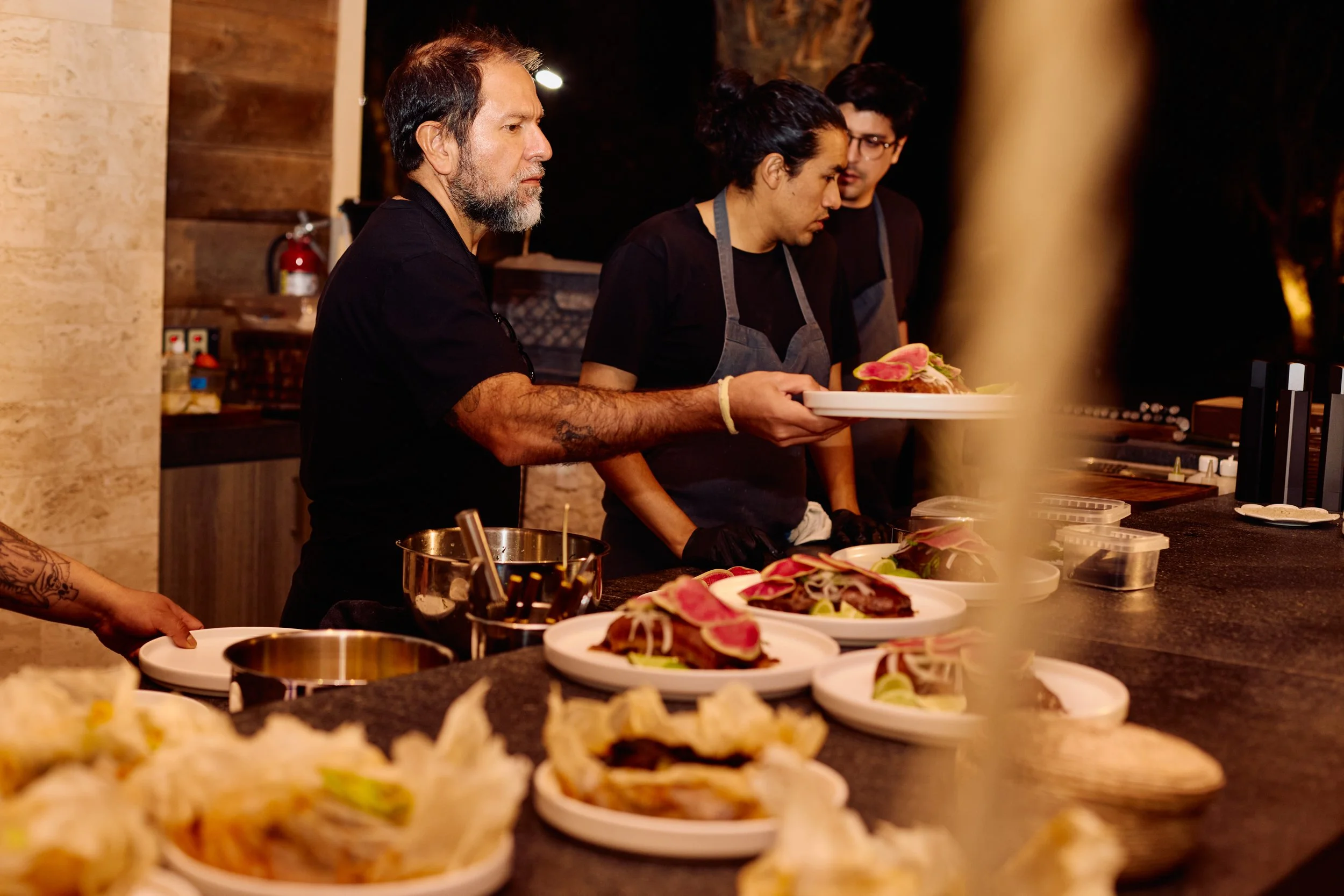 Three chefs, two women and one man, preparing dishes with slices of raw meat and garnishes on plates at a restaurant counter.