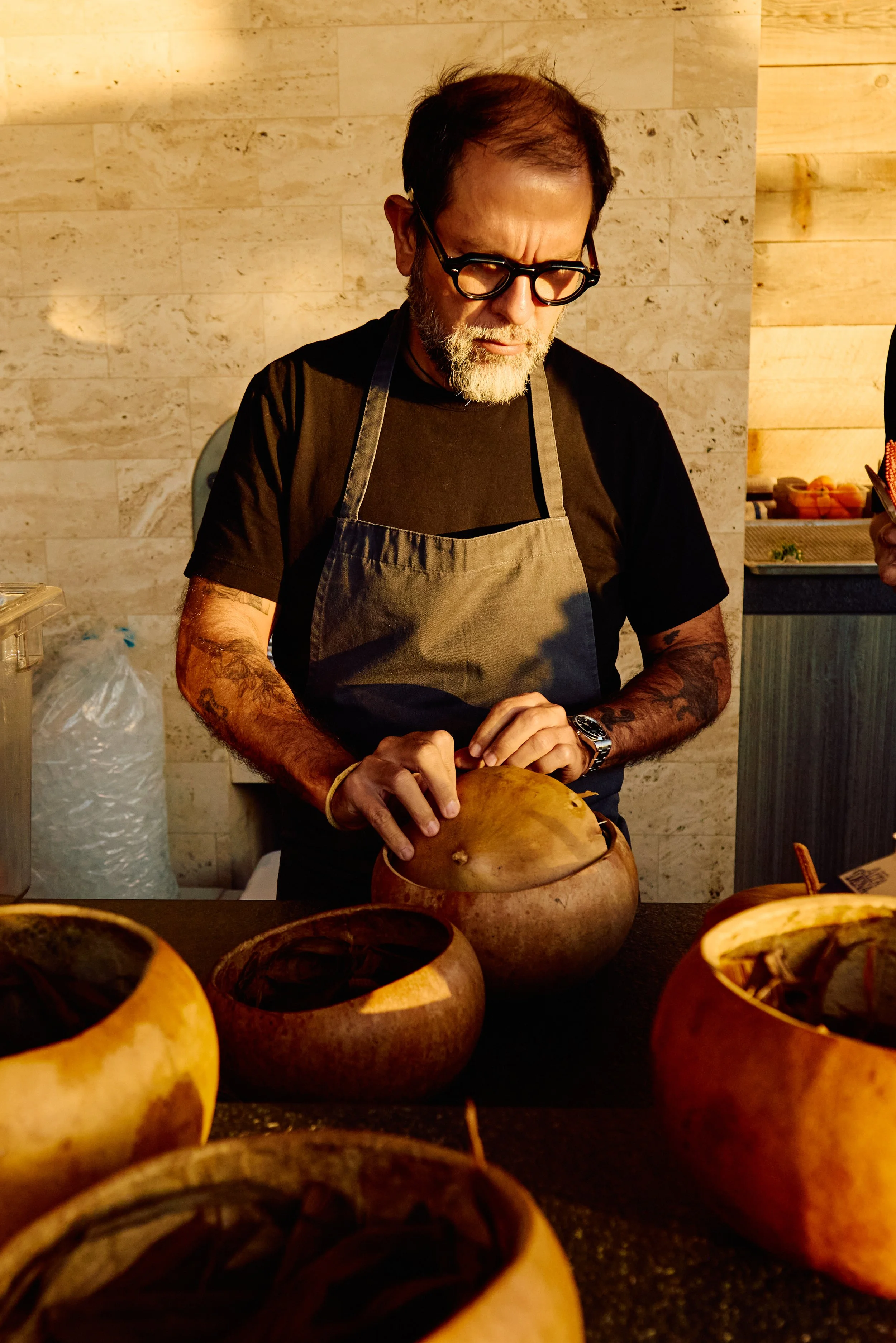 A man with glasses and a beard, wearing a black shirt and apron, is carving a pumpkin at a table with other carved pumpkins in front of him.