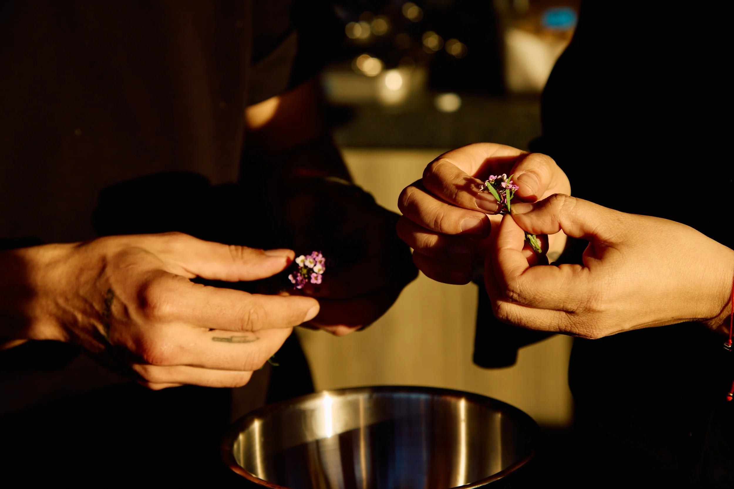 Two people exchanging flowers over a metallic bowl, with focus on their hands, in a warm, dimly lit setting.