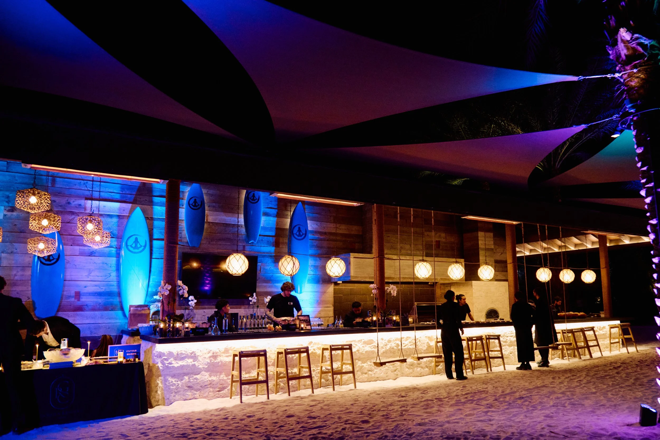 Beachside bar at night with surfboard decorations, hanging lanterns, and a few people standing or sitting near the counter under colorful lighting.