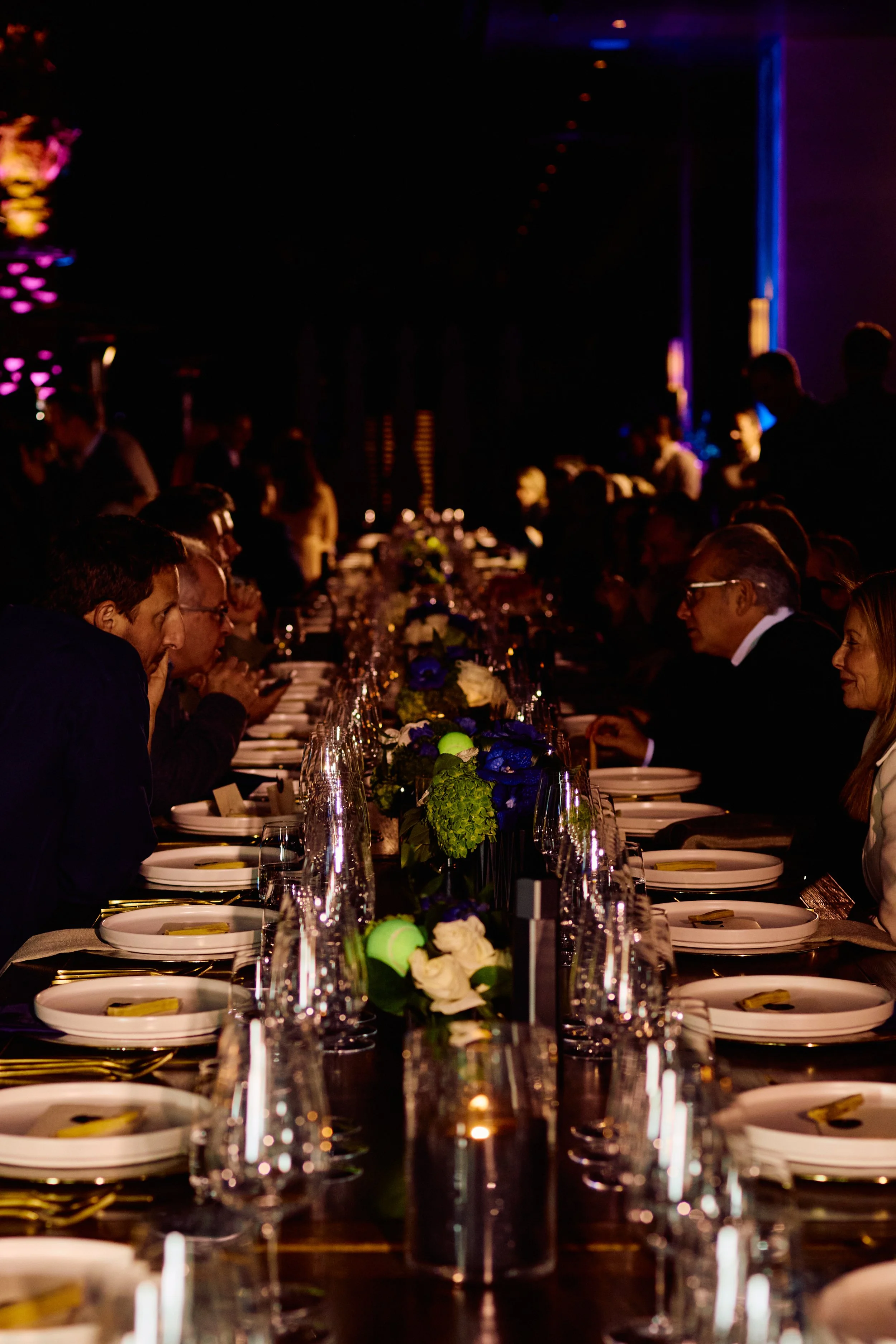 A long banquet table set for a formal event with white plates, gold utensils, wine glasses, and flower arrangements with white, blue, and green flowers, in a dimly lit room.