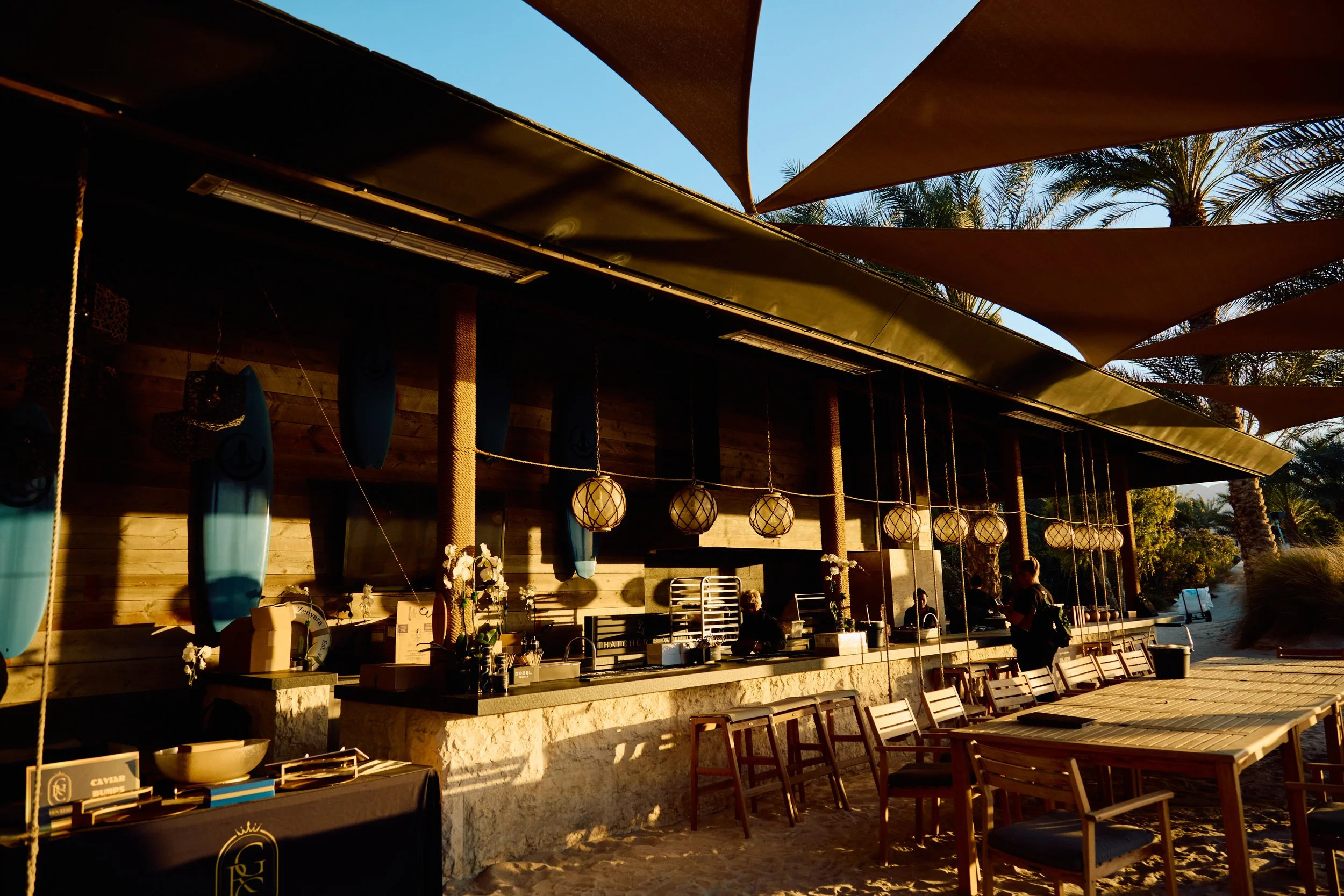 Beachside outdoor bar area with surfboards hanging on the wall, round paper lanterns, wooden tables and chairs, and palm trees in the background during sunset.