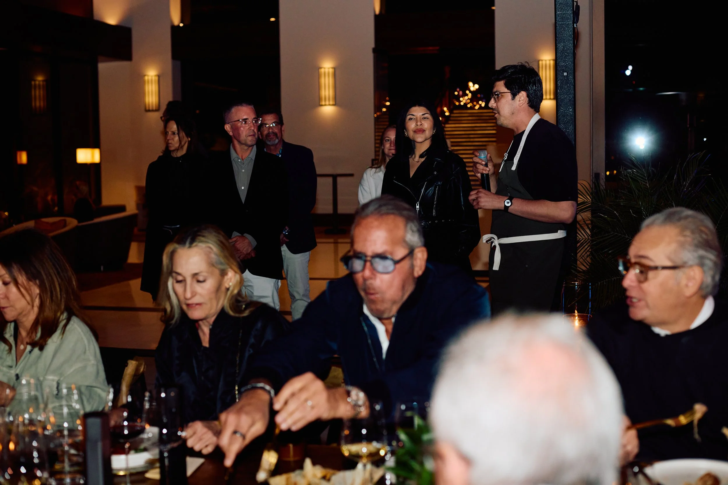 People gathered at a dining table and a woman speaking into a microphone at an event in a warmly lit indoor space.