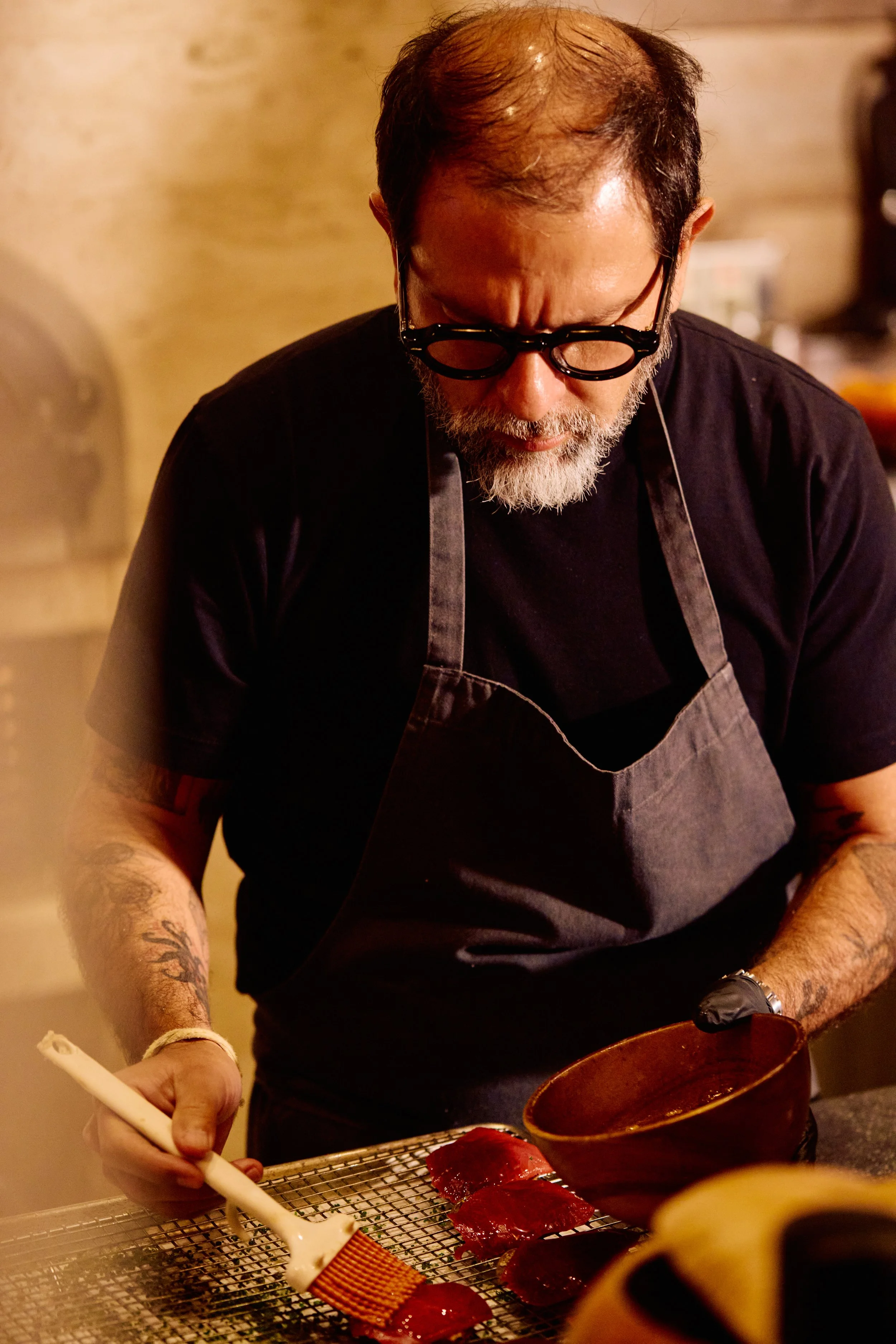 A man with glasses and a gray beard using a brush to glaze slices of meat on a rack in a kitchen.