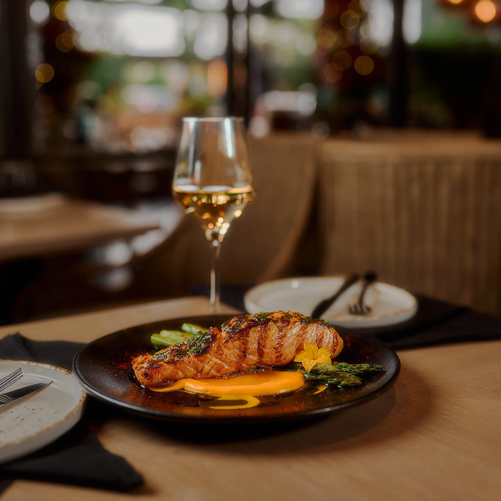 A plate of grilled salmon with asparagus and sauce on a black plate, a glass of white wine, and empty plates and utensils on a wooden table in a restaurant setting.
