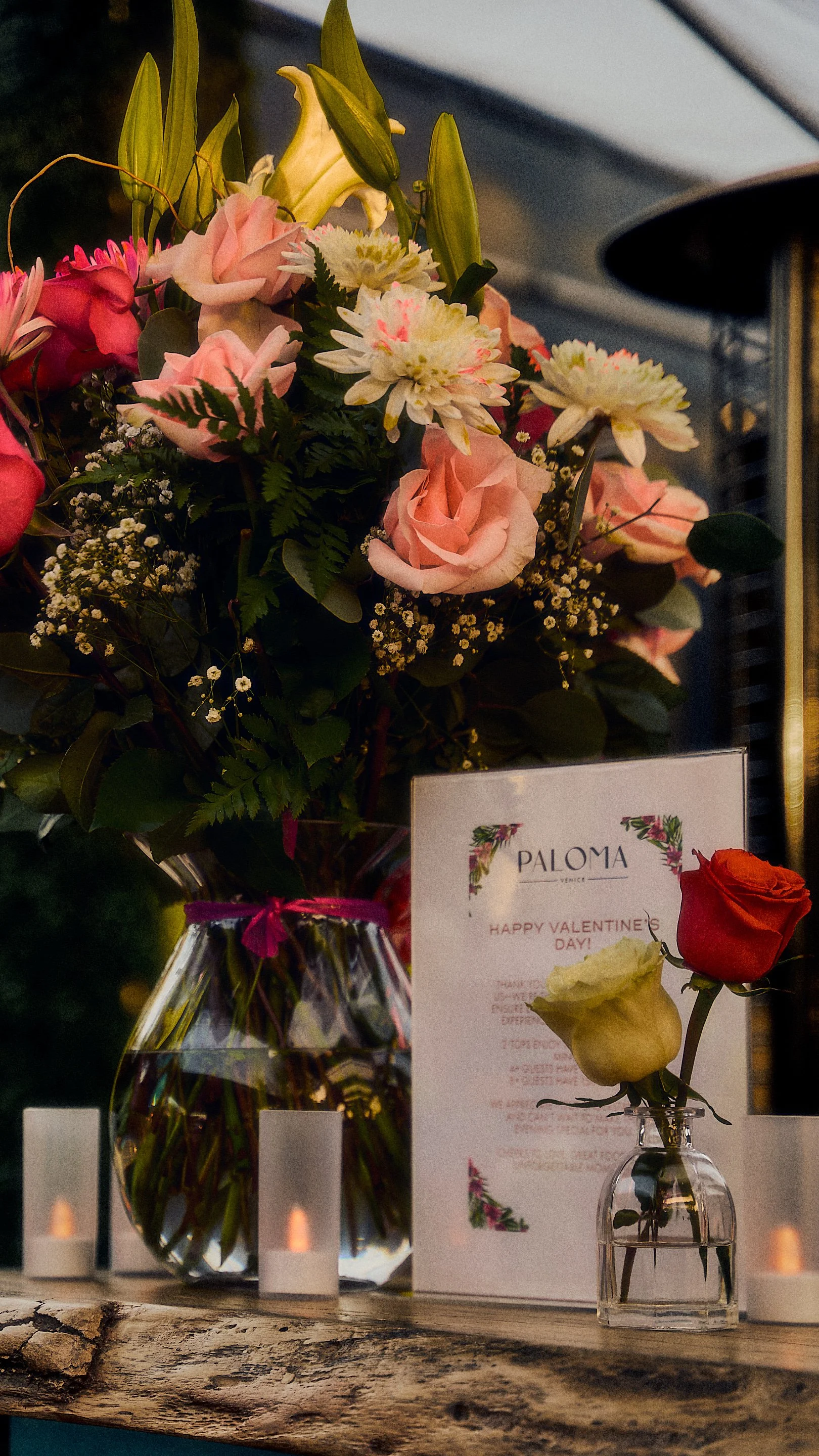 A floral arrangement of pink and white flowers in a glass vase, with a romantic Valentine's Day card and two small flower vases with roses, on a wooden surface with candles