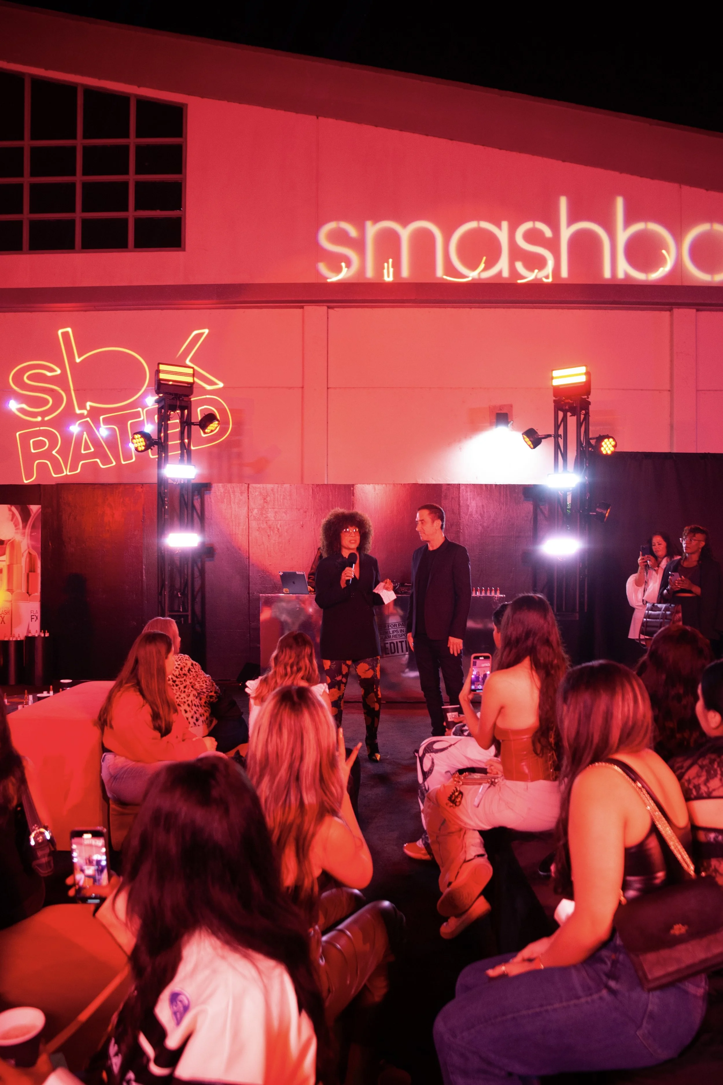 At an indoor event with red lighting, a woman with curly hair and glasses is speaking into a microphone. A man stands beside her, holding a paper. Audience members are seated and taking photos. Neon signs reading 'smashbox' and 'skbratd' are visible