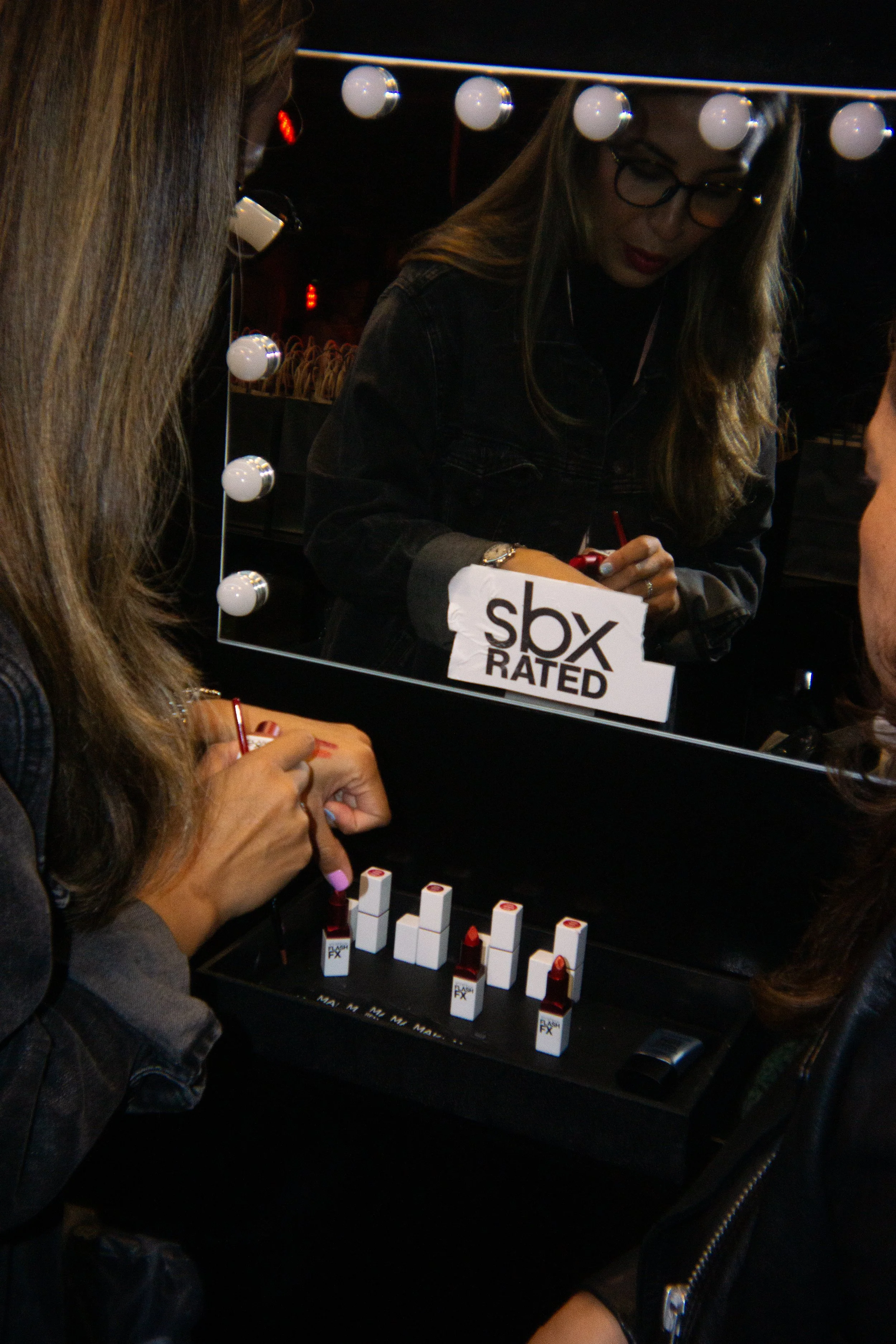 A woman applies lipstick in front of a mirror surrounded by lights, with a sign reading 'SBO Rated' in the reflection, and lipstick tubes on a black tray.
