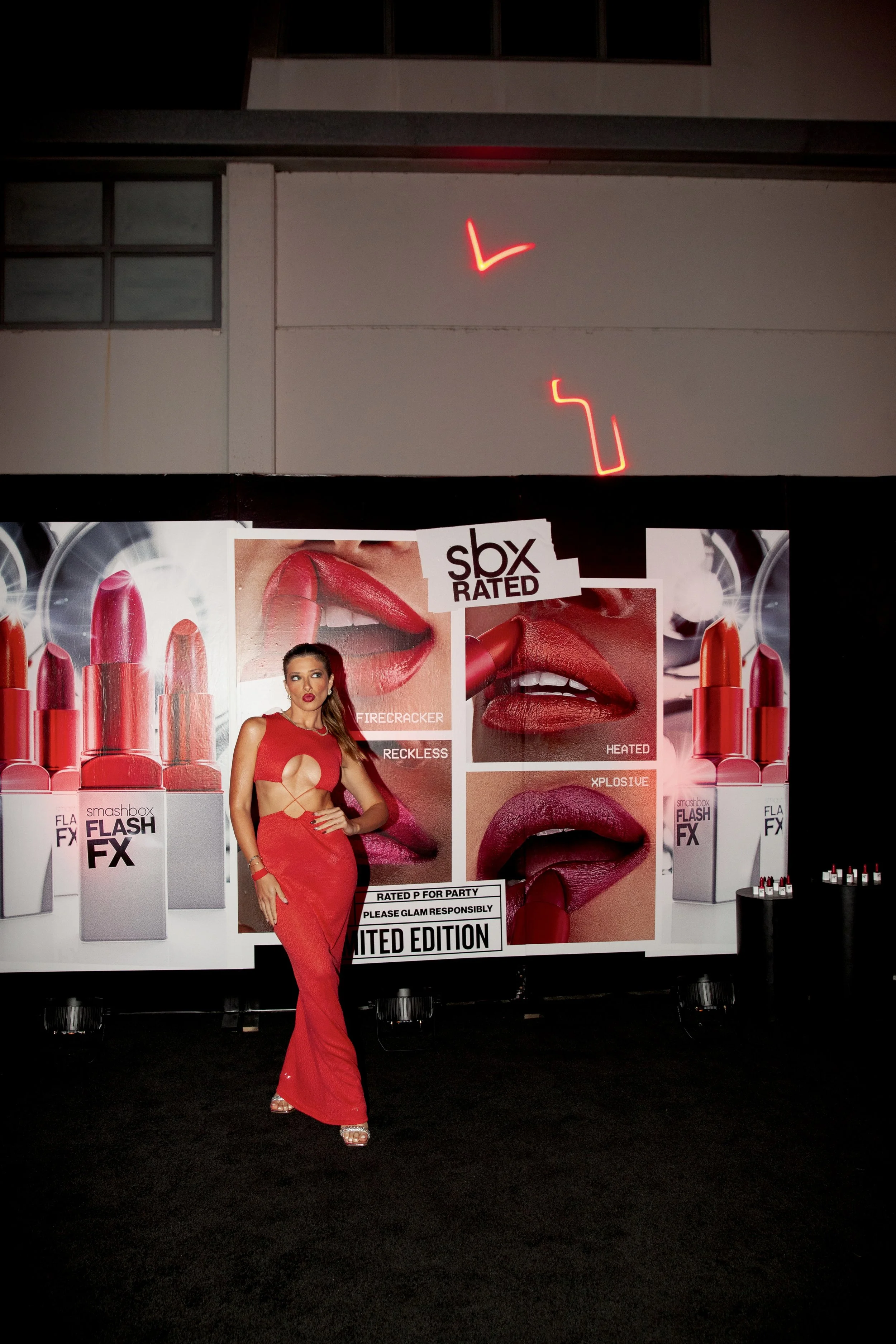 A woman in a red dress standing in front of a large advertisement for lipstick, with close-up images of lips and lipstick tubes, at a promotional event. Neon lights are visible above the display.