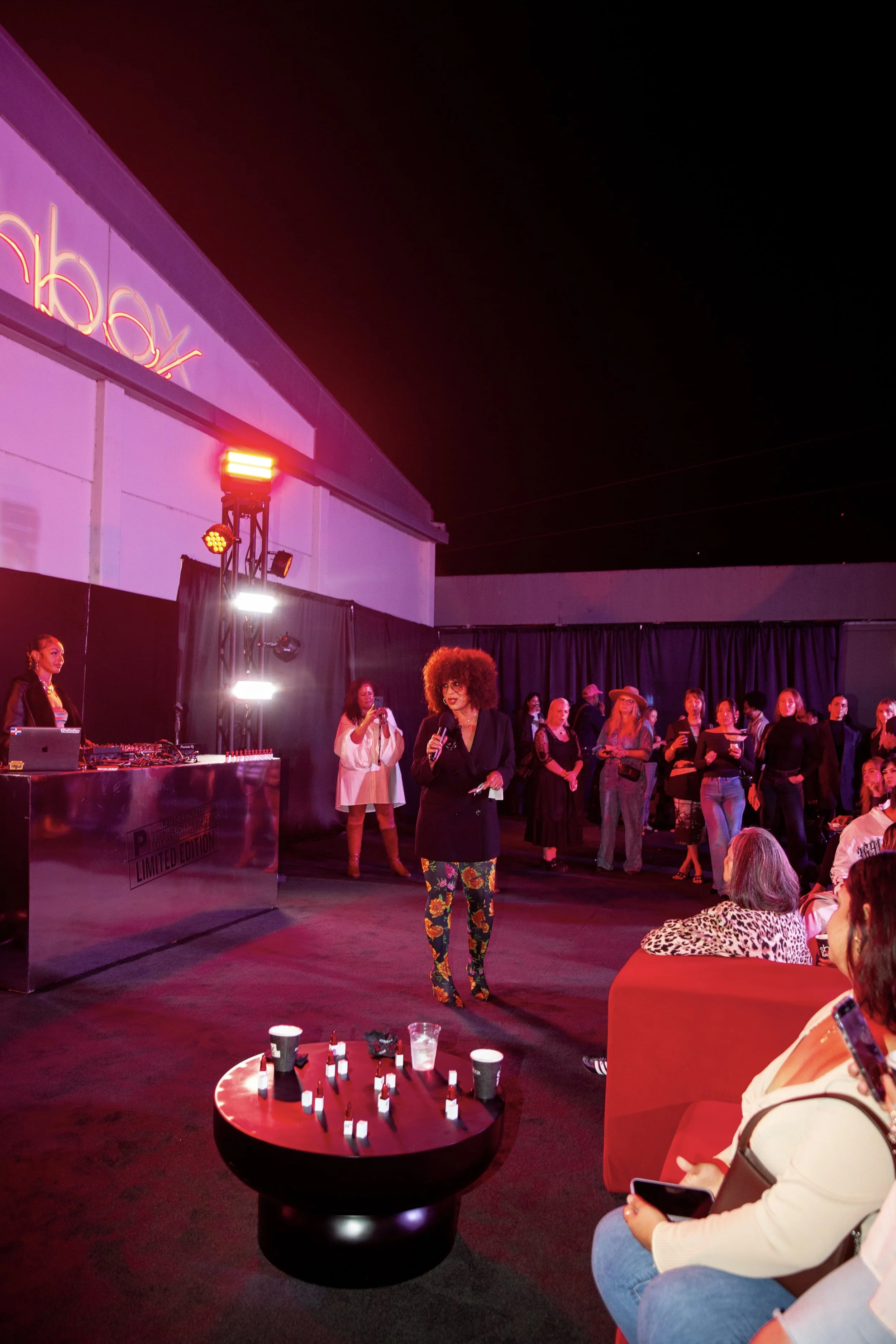 A woman with curly hair and floral pants speaking into a microphone at a lively indoor event with people watching and a DJ setup nearby.