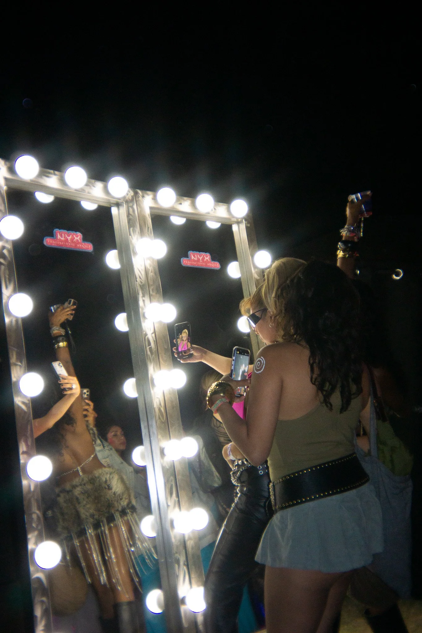 Group of women taking photos of themselves in front of a large illuminated mirror at a party or club event.