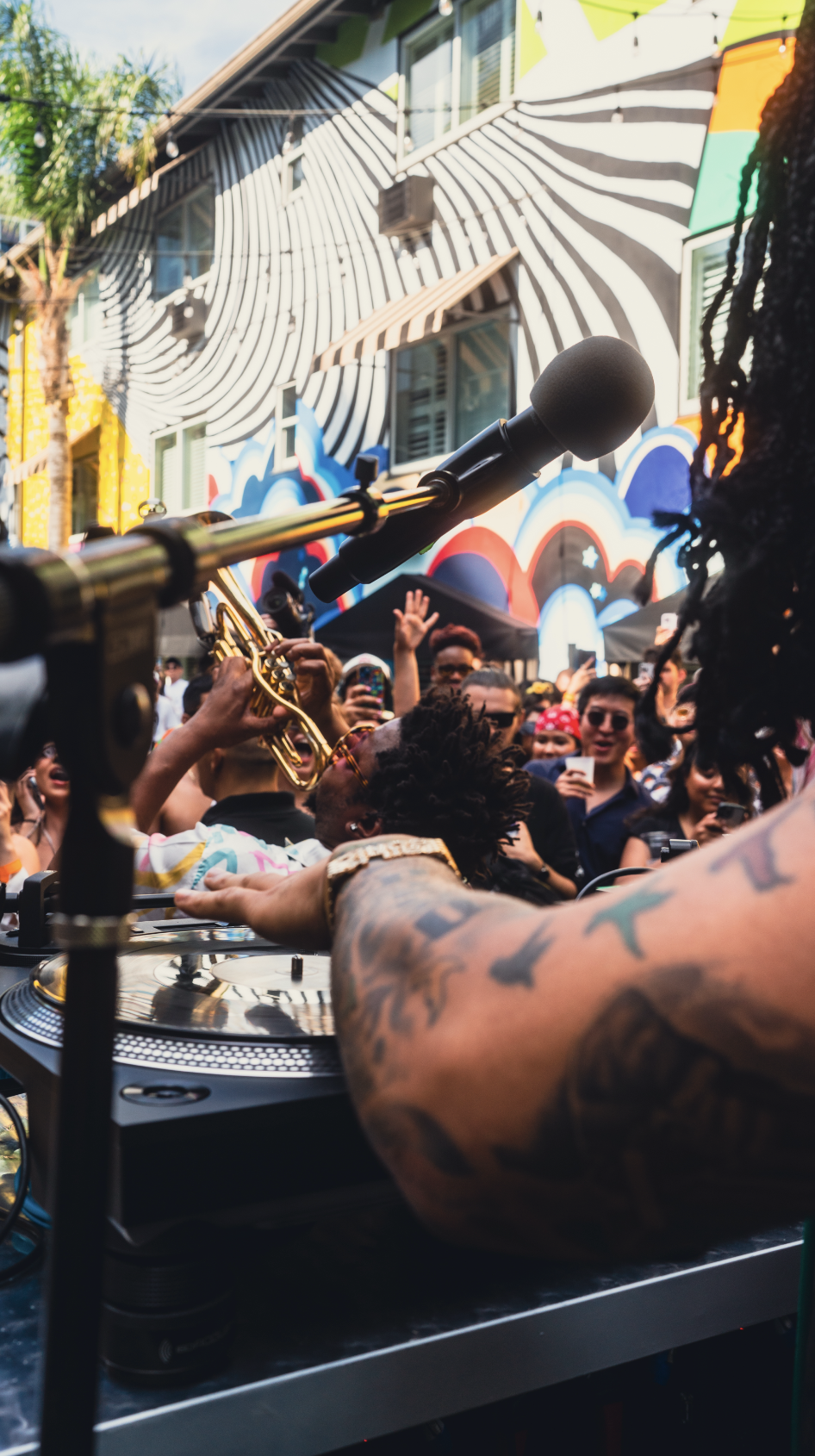 A DJ performs at an outdoor event with a crowd of people dancing and taking photos, against a colorful mural on a building.