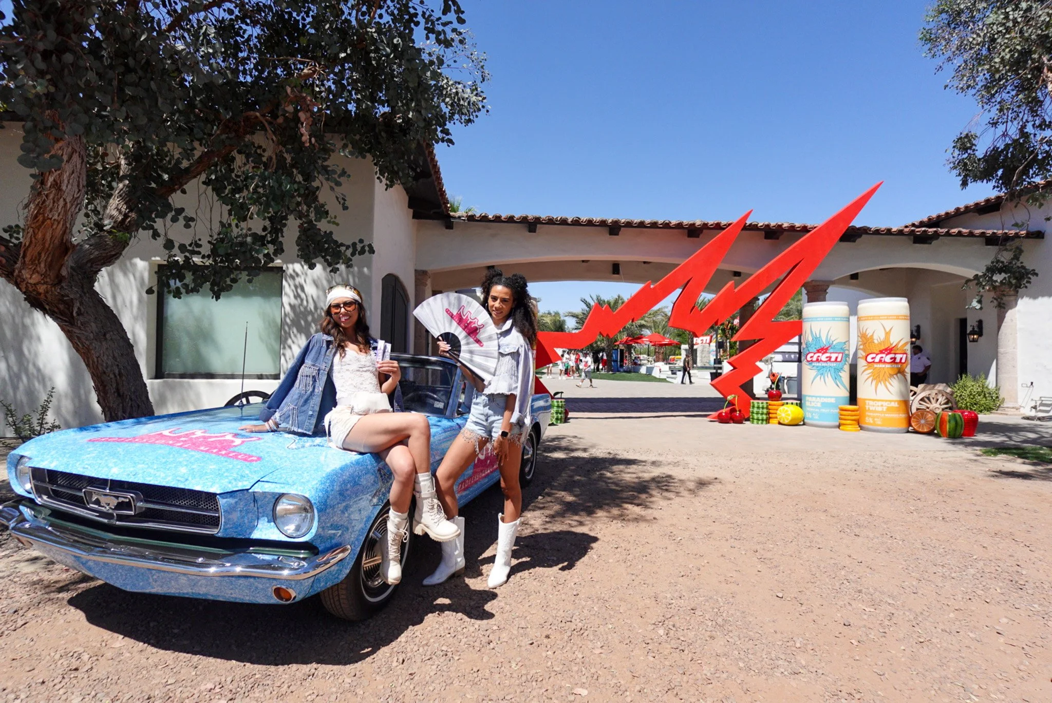 Two young women sitting on a vintage blue car, smiling and posing for the camera, with a large red lightning bolt decoration behind them. The background features a white building with a courtyard, palm trees, and two large cylindrical objects with th