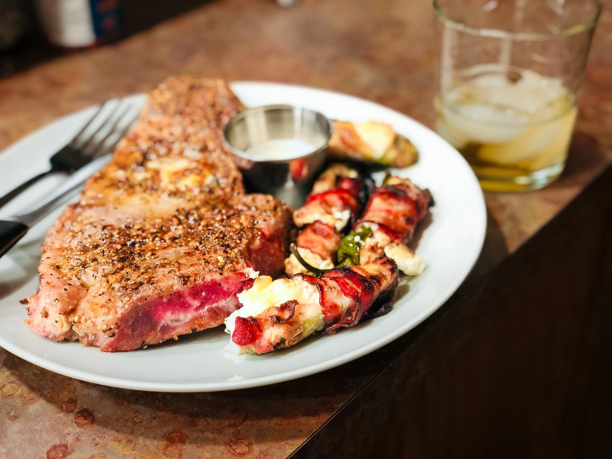 A plate of cooked steak, bacon-wrapped jalapenos with cream cheese, a small cup of dipping sauce, and a glass of iced beverage with lemon slices.