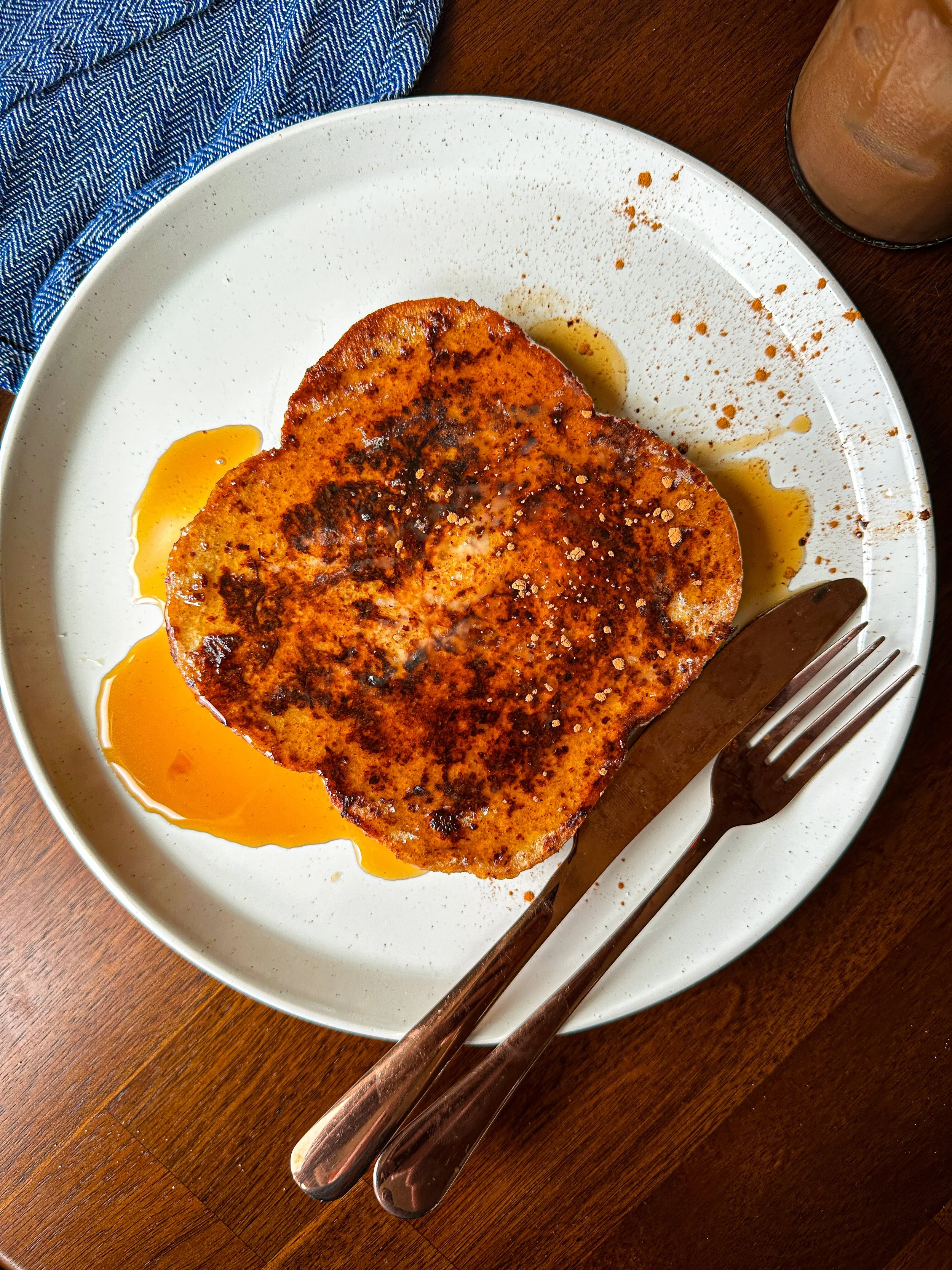 A plate with a cooked piece of French toast topped with syrup, salt, and possibly cinnamon. There is a fork and knife on the plate, and a glass of iced beverage in the corner.