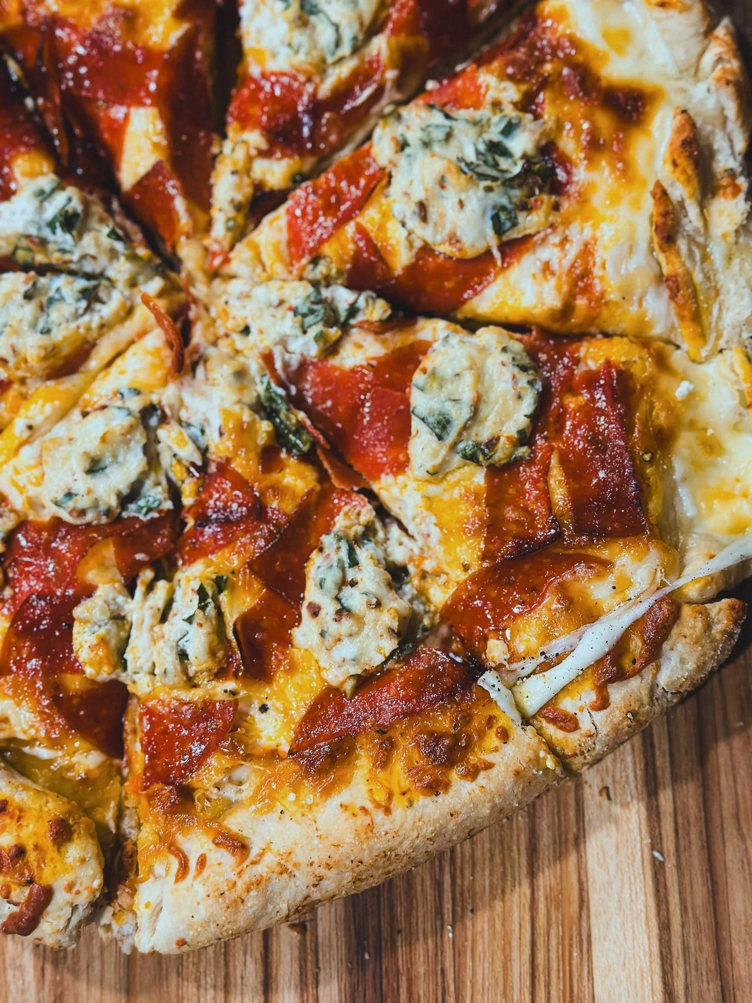 Close-up of a pizza with cheese, pepperoni, chicken, and herbs on a wooden surface.