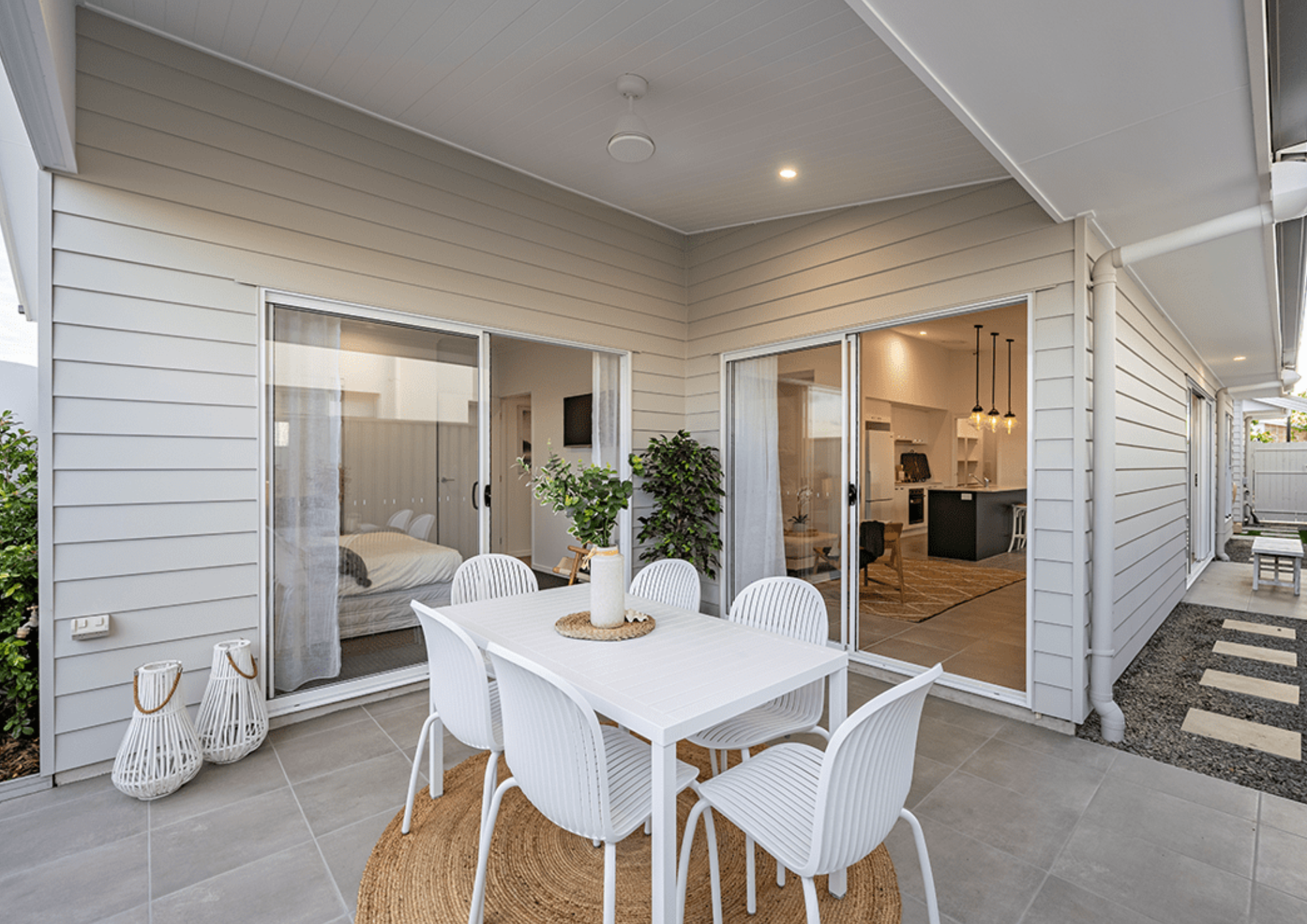 Outdoor patio with white dining table and six white chairs, with a vase and greenery on top, in front of a beige house with sliding glass doors revealing an interior with a bedroom and kitchen area.