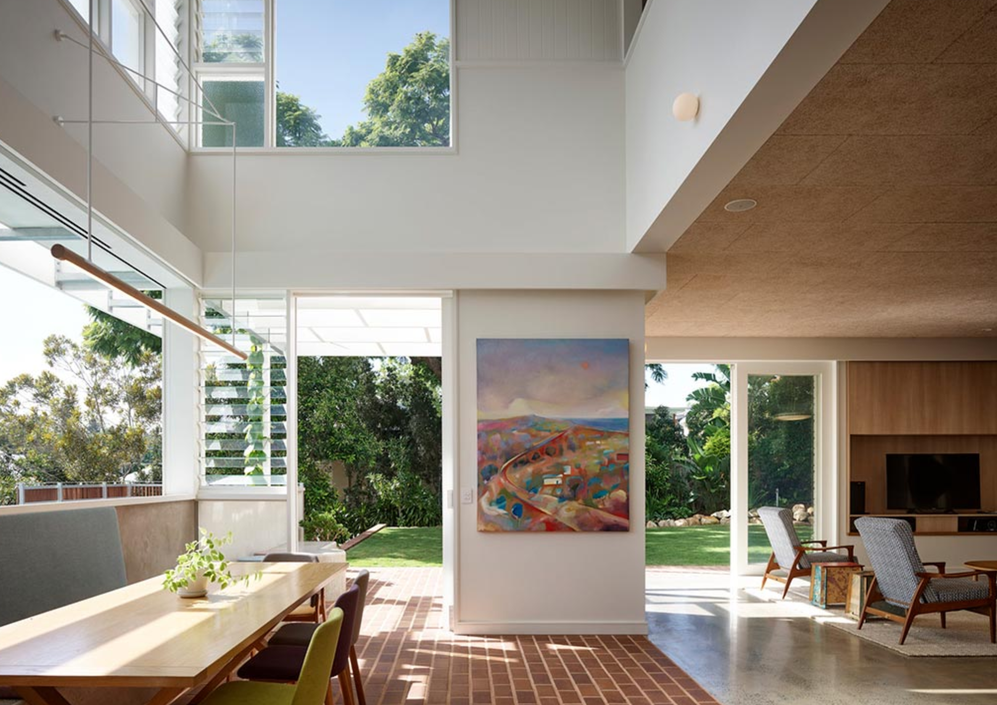 Modern living room with large windows, greenery outside, a colorful landscape painting on the wall, and seating area with chairs.