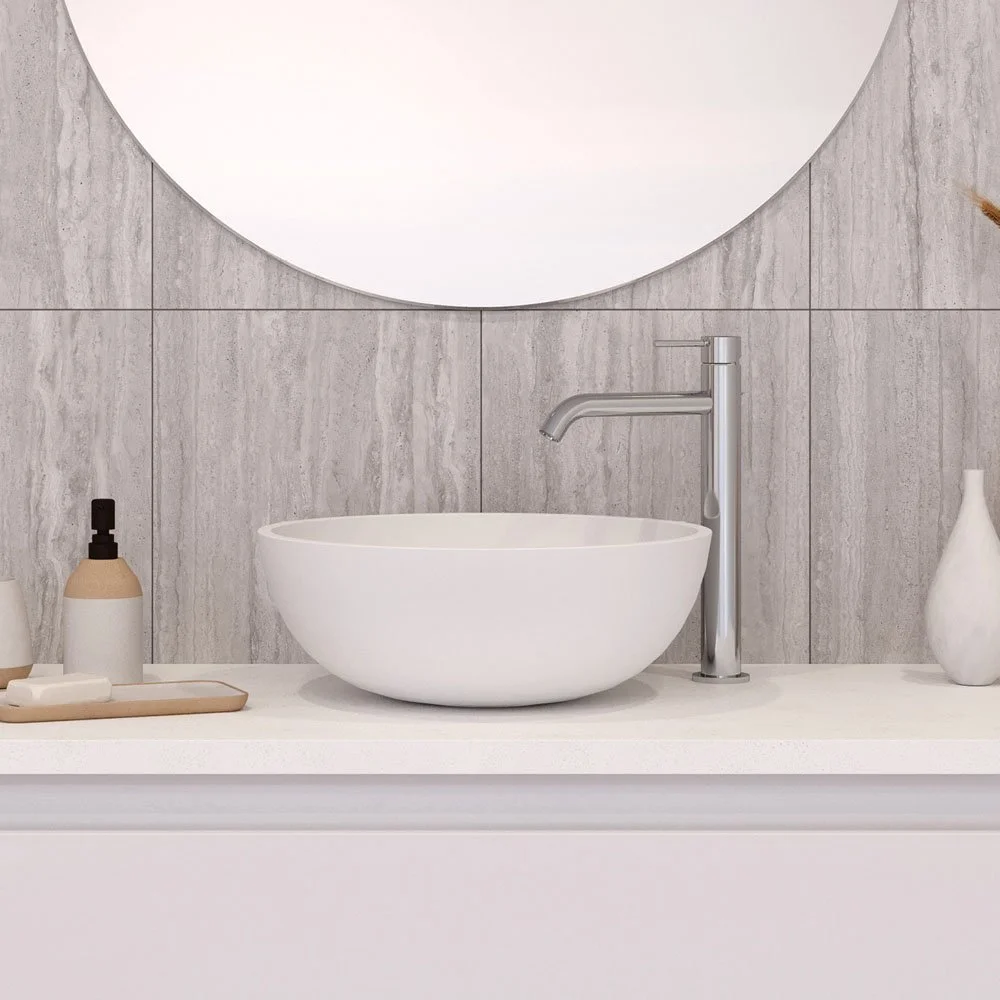 A modern bathroom sink area with a white vessel sink, a silver faucet, and decorative bottles on a white countertop, with a large round mirror reflecting the wall behind.