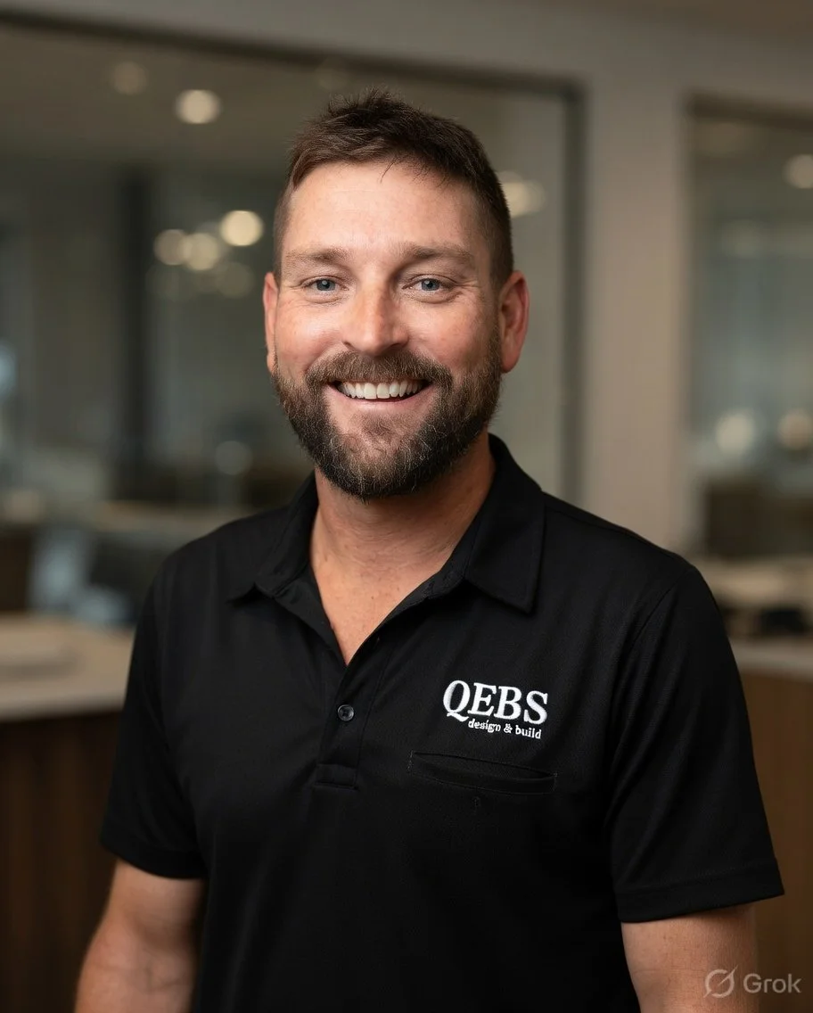 A smiling man with a beard and short brown hair, wearing a black CEO polo shirt, standing in an office environment.