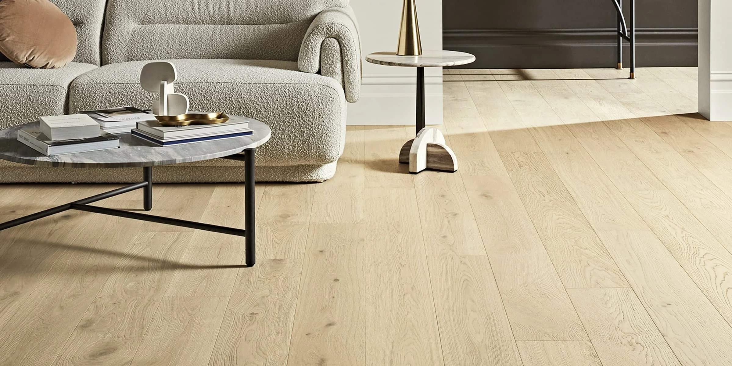 Living room with a beige textured sofa, a round marble coffee table with black metal legs holding stacked books and a white sculpture, and a small side table with a black metal stand and a white marble top, all on light hardwood flooring.
