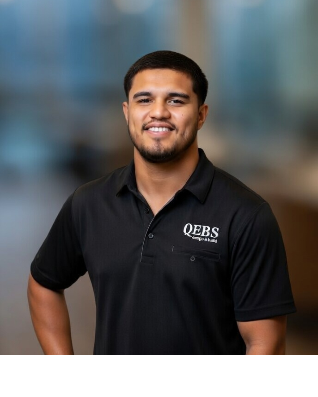 Portrait of a young man with short black hair and a beard, wearing a black polo shirt with the logo 'QEB S' on it, smiling and standing with one hand on his hip against a blurred background.