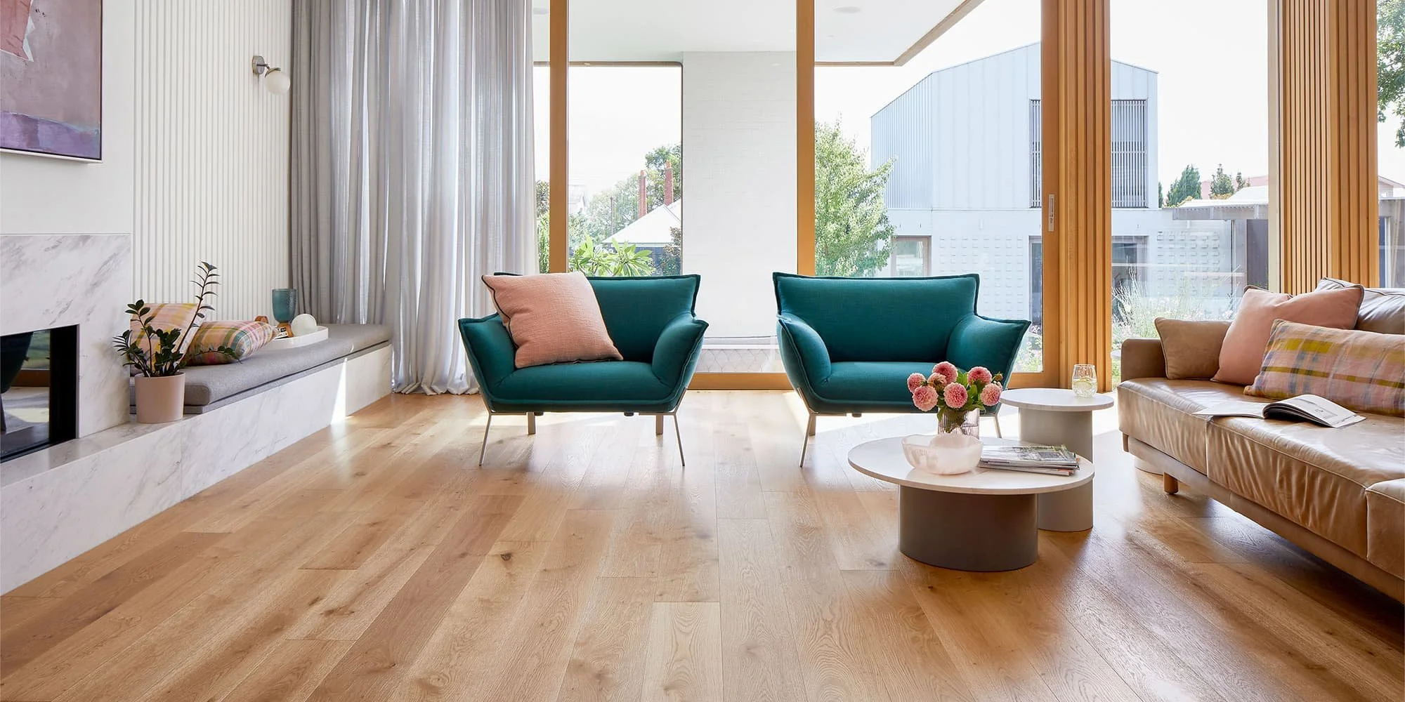 Modern living room with wood flooring, teal armchairs with pink pillows, a beige sofa with pink cushions, white round coffee tables with a vase of pink flowers and magazines, large windows with curtains, and a fireplace with plants on the mantel.