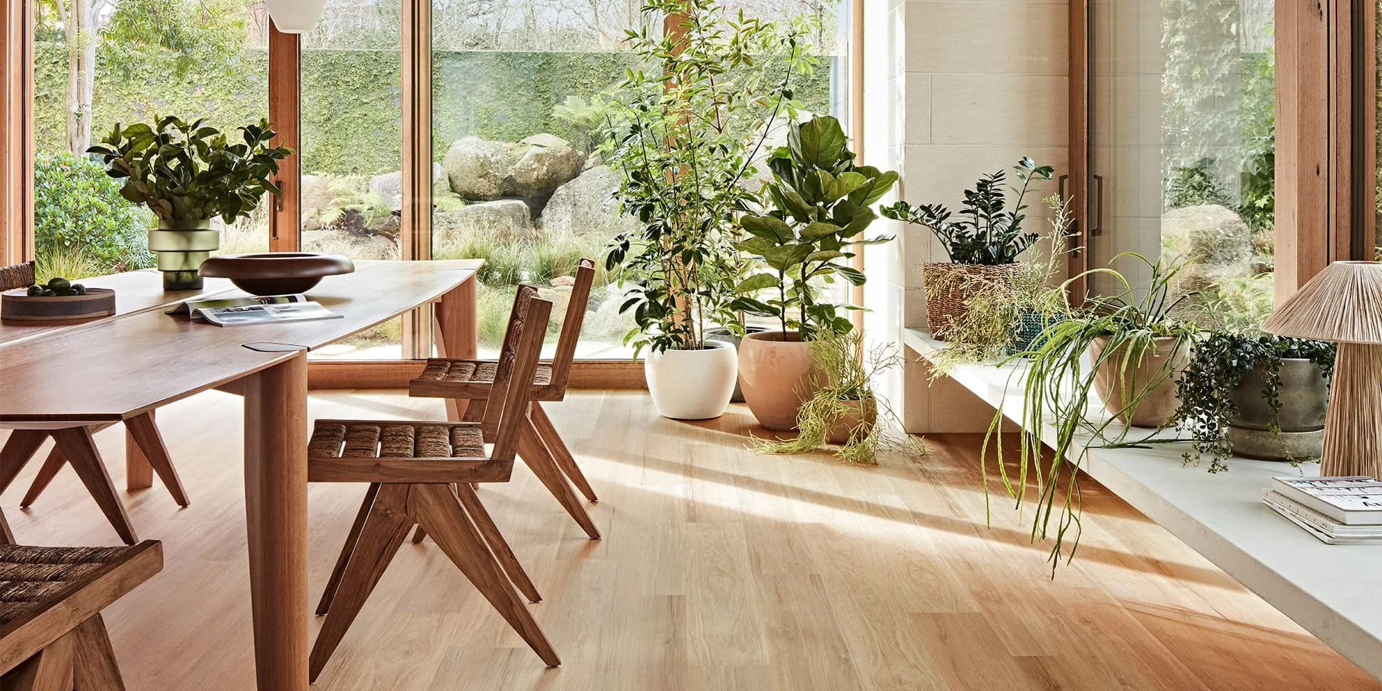 A dining room with a wooden table, wooden chairs, and a large window revealing a garden with rocks and bushes. Potted plants are on a ledge and on the floor near the window.