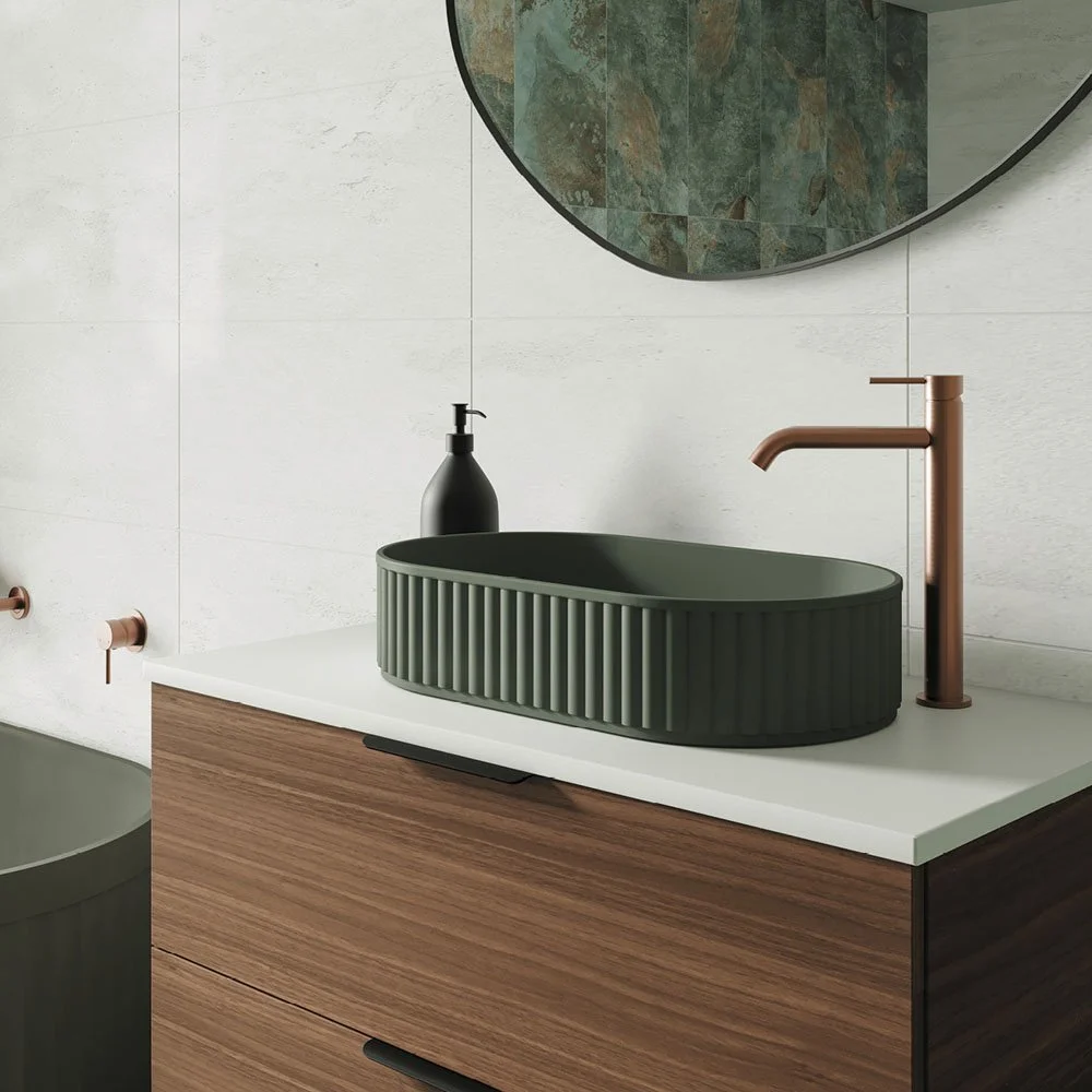 A modern bathroom sink setup featuring a green, ribbed oval vessel sink on a white countertop, a copper faucet, and a black soap dispenser. There is a round mirror above the sink and a white tiled wall in the background.