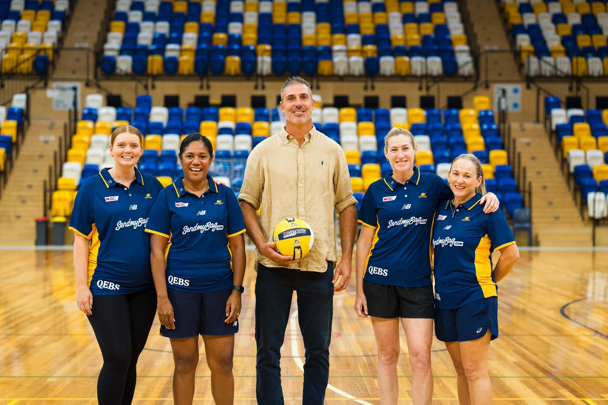 Group of five people, three women and one man, standing on a sports court in an indoor stadium, smiling, with empty yellow, blue, and white stadium seats in the background, the man holding a volleyball.