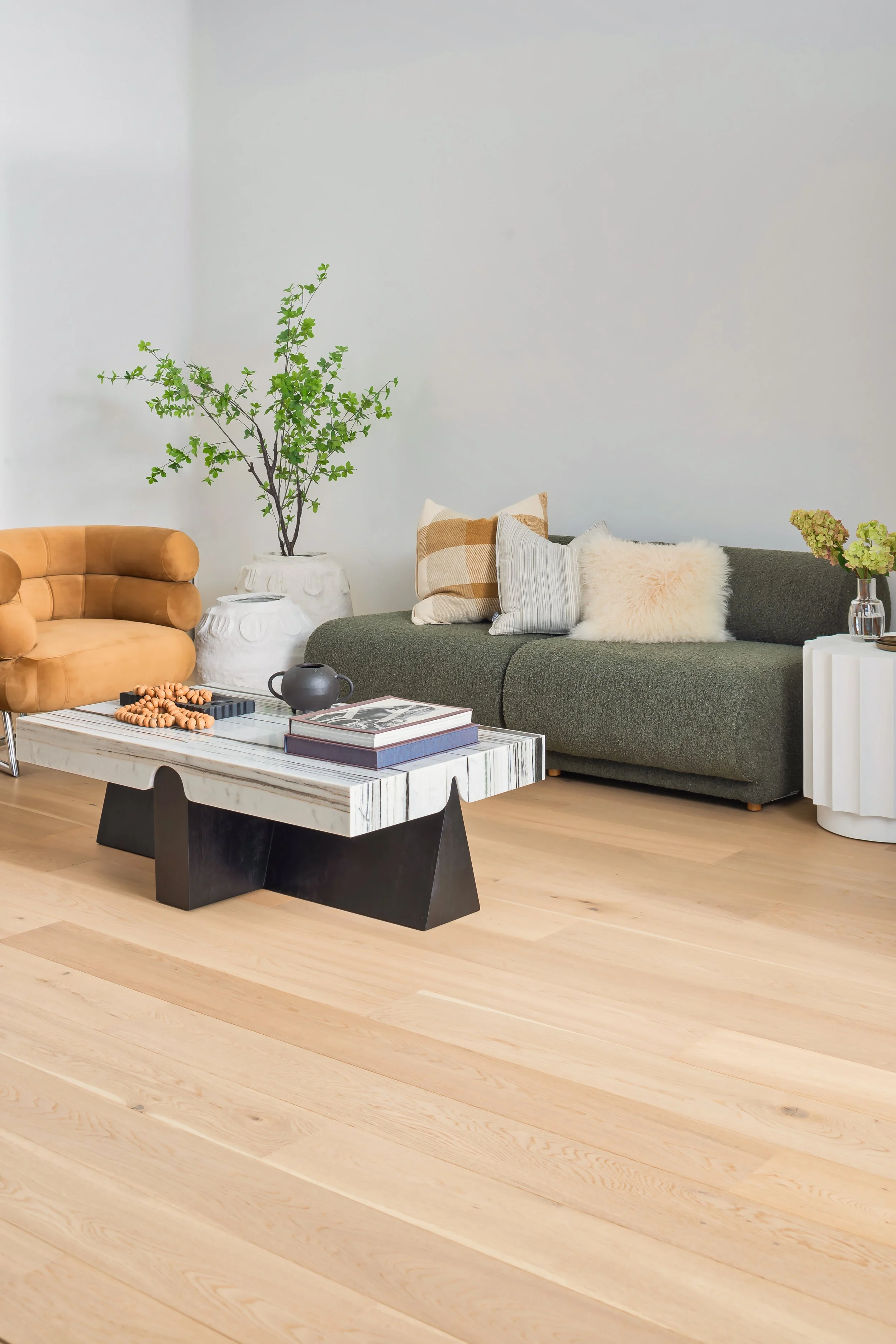 A modern living room with a beige armchair, a dark green sofa with pillows, a white marble coffee table with books and decor, a large potted plant, and wooden flooring.