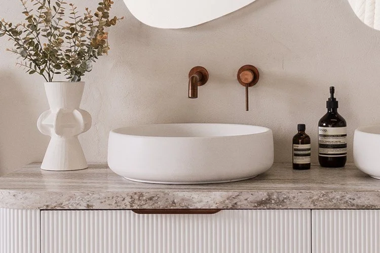 Bathroom countertop with a white vase holding green foliage, a round vessel sink, and three bottles of soap or lotion, with a round mirror above.