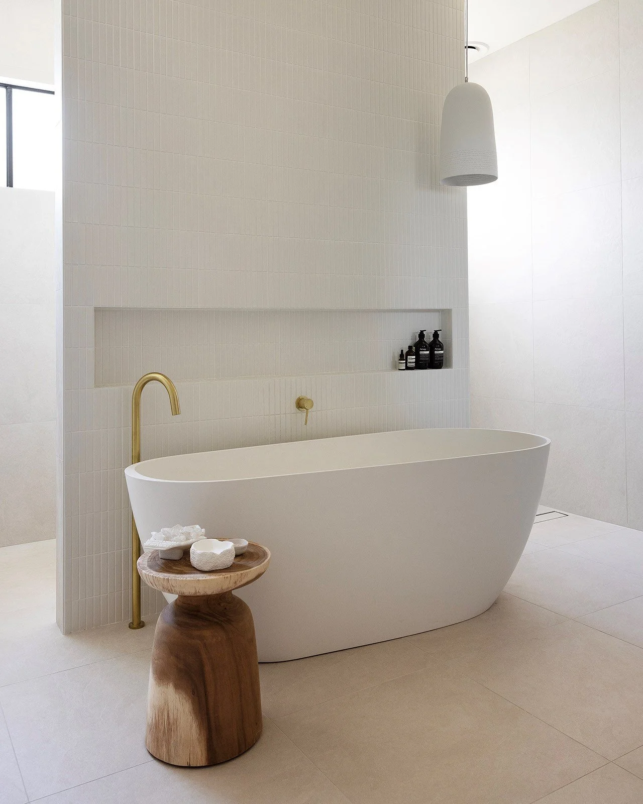Minimalist bathroom with a white freestanding bathtub, a gold faucet, a small wooden side table with decorative stones, and black bottles on a built-in shelf. White tiles and a large white pendant light.