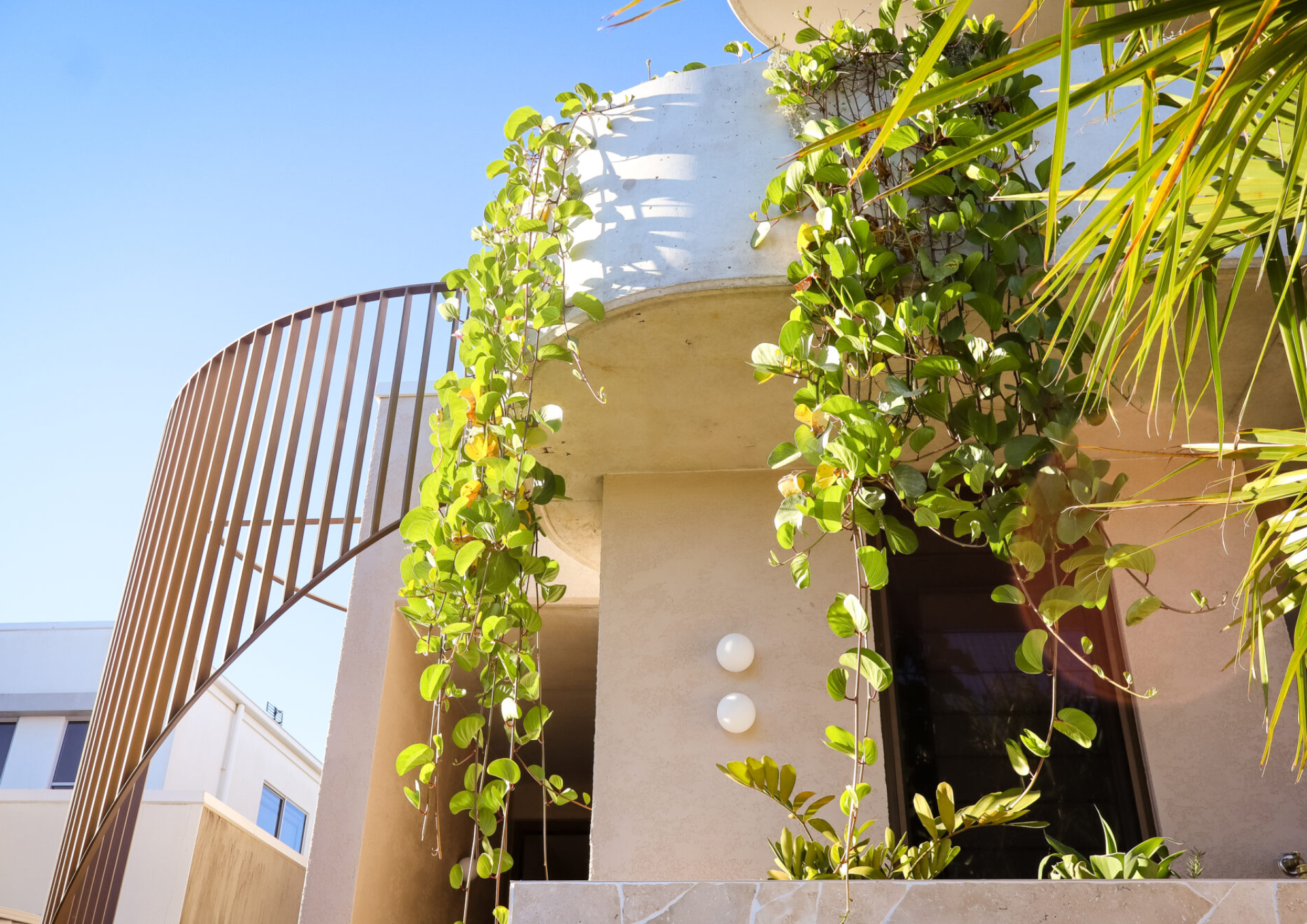 A modern building with a rounded balcony and a metal railing, decorated with green vines and plants, against a clear blue sky.