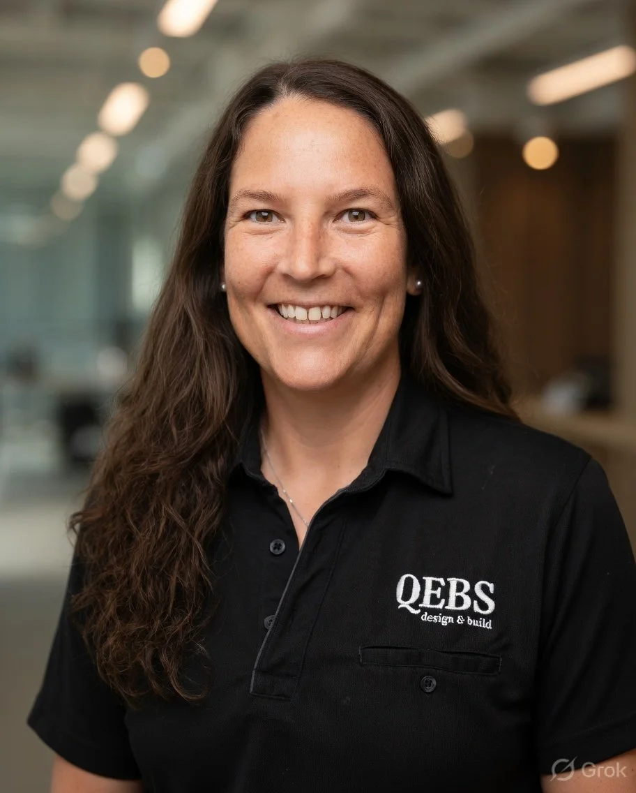 Smiling woman with long brown hair wearing a black polo shirt with 'QERS design & build' logo, standing in an indoor office setting.