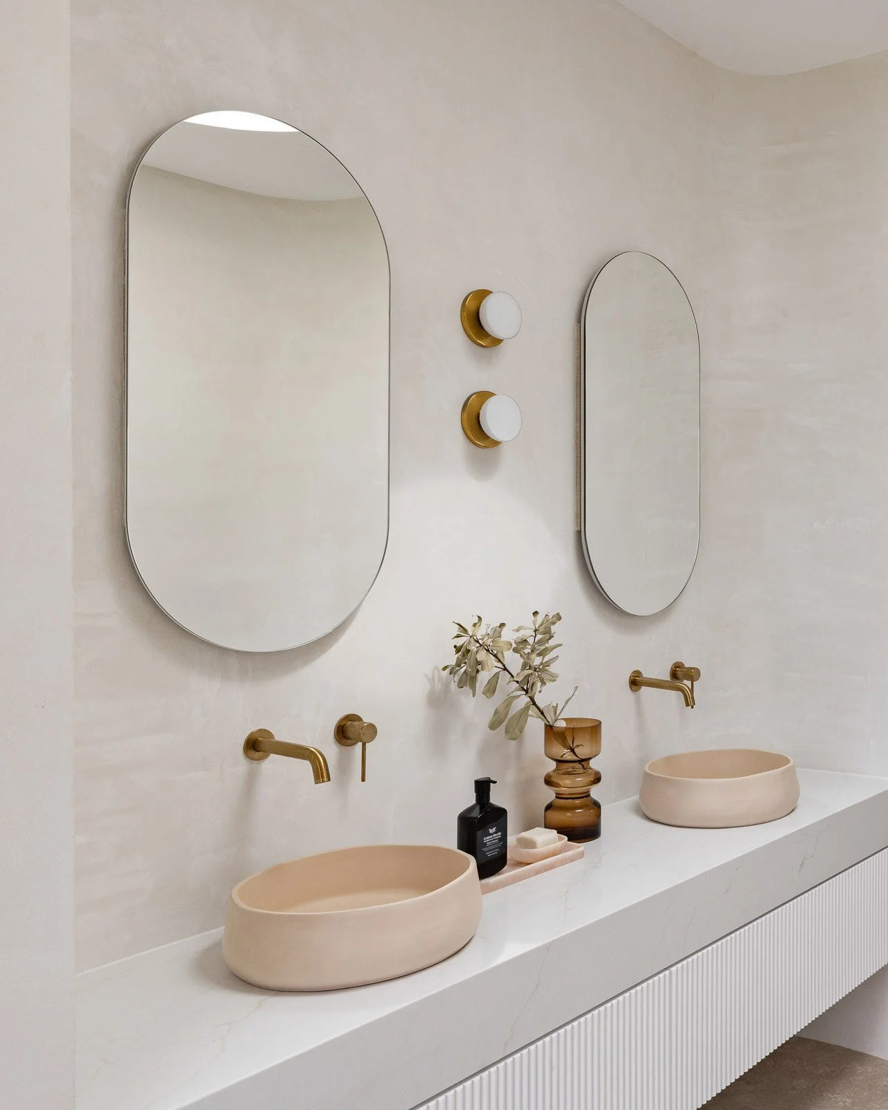 Modern bathroom vanity with two oval mirrors, two beige vessel sinks with gold faucets, two white wall-mounted lights, and decor including a black soap dispenser, a small beige dish, and a brown vase with green foliage.