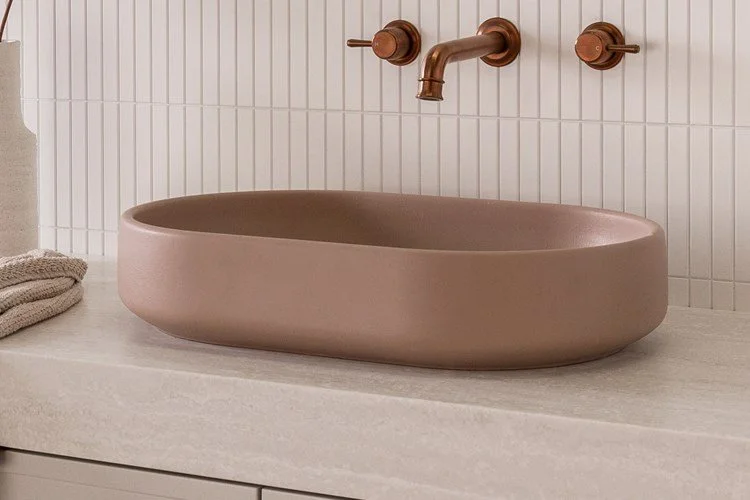 A modern bathroom sink with a pinkish-beige oval basin, bronze faucet, and wall-mounted handles, set against white tiled wall.