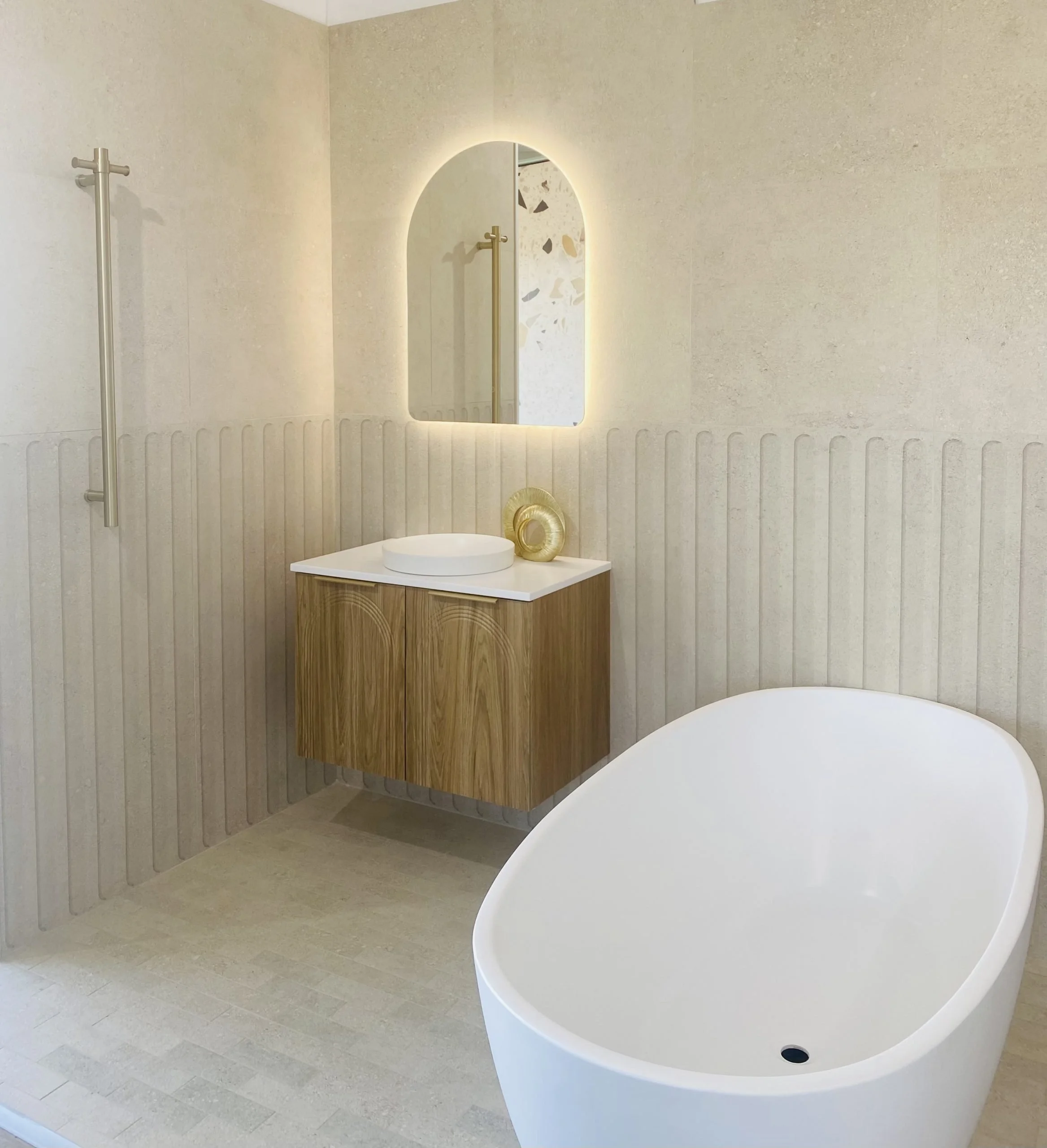 Modern bathtub and vanity in a minimalist bathroom with beige textured walls, a wooden cabinet, and a backlit mirror.